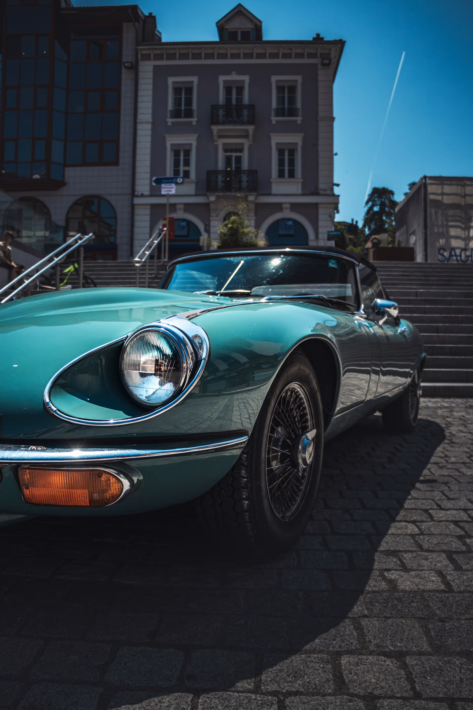 Front side view of a turquoise vintage convertible car parked on cobblestone street with a building and stairs in the background.