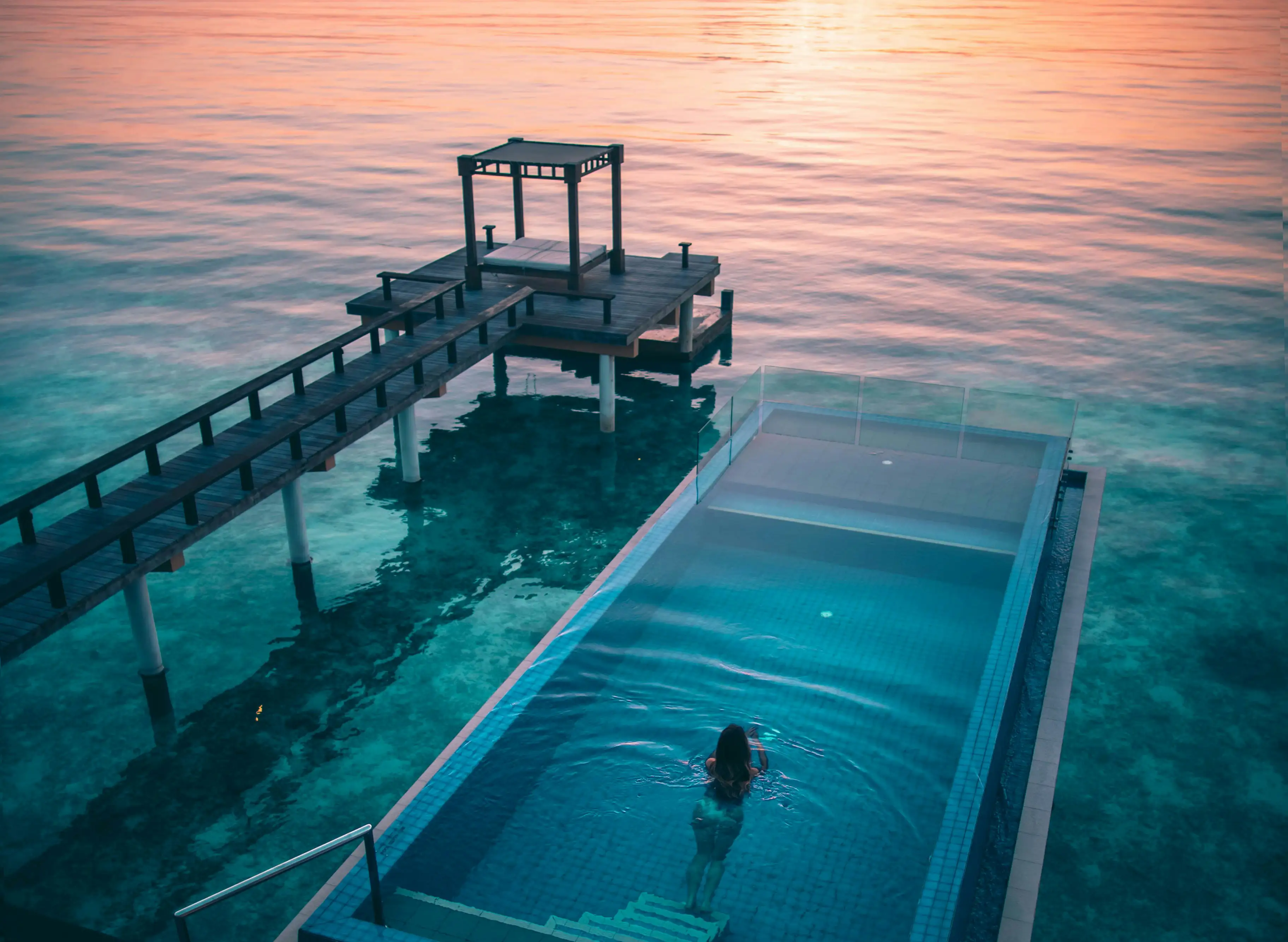 Woman swimming in an infinity pool overlooking clear ocean water at sunset with a wooden pier and gazebo.
