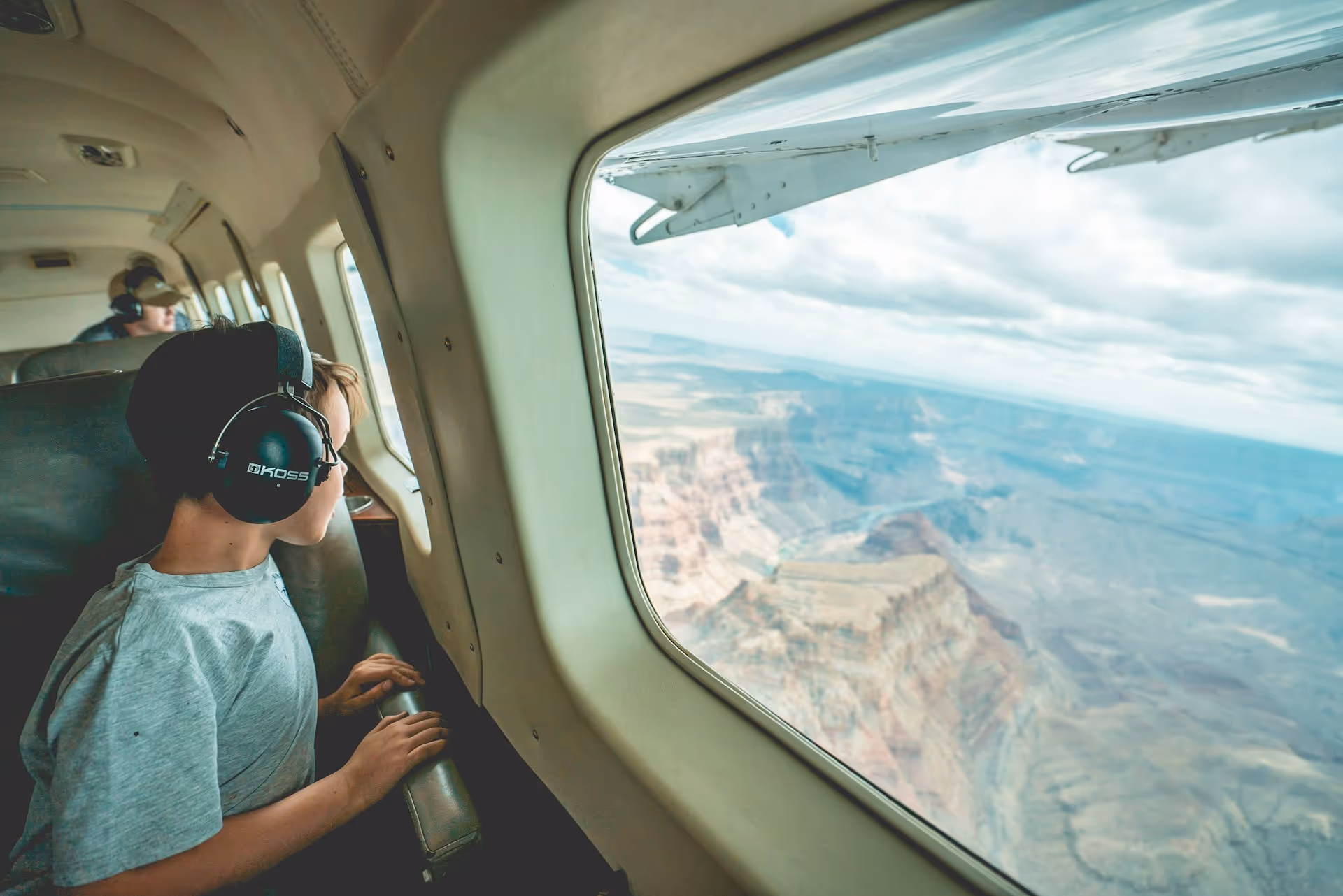 Boy wearing headphones looking out airplane window at canyon landscape below.