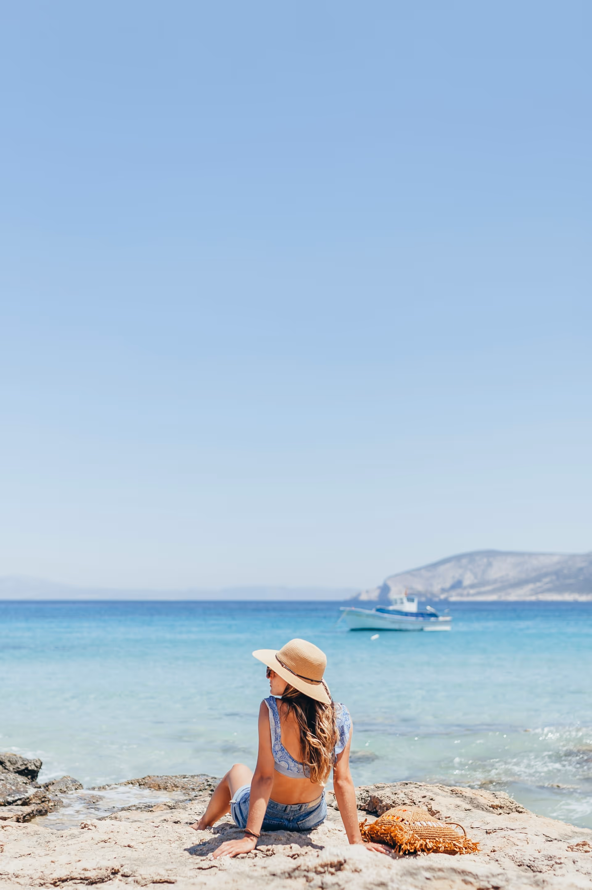 Woman in a sunhat and blue top sitting on a rocky shore looking at a boat on clear turquoise sea with distant mountains.