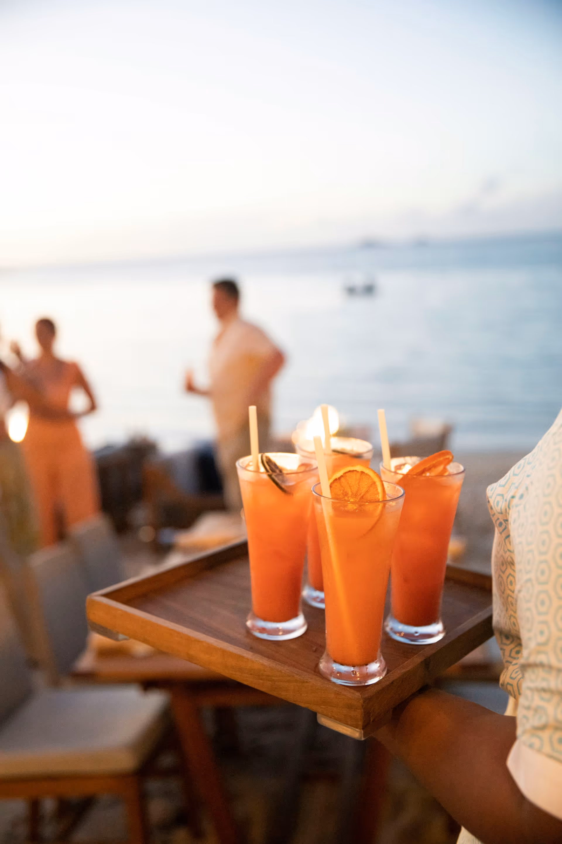 Serving tray with four orange cocktails garnished with citrus slices at a beachside gathering during sunset.