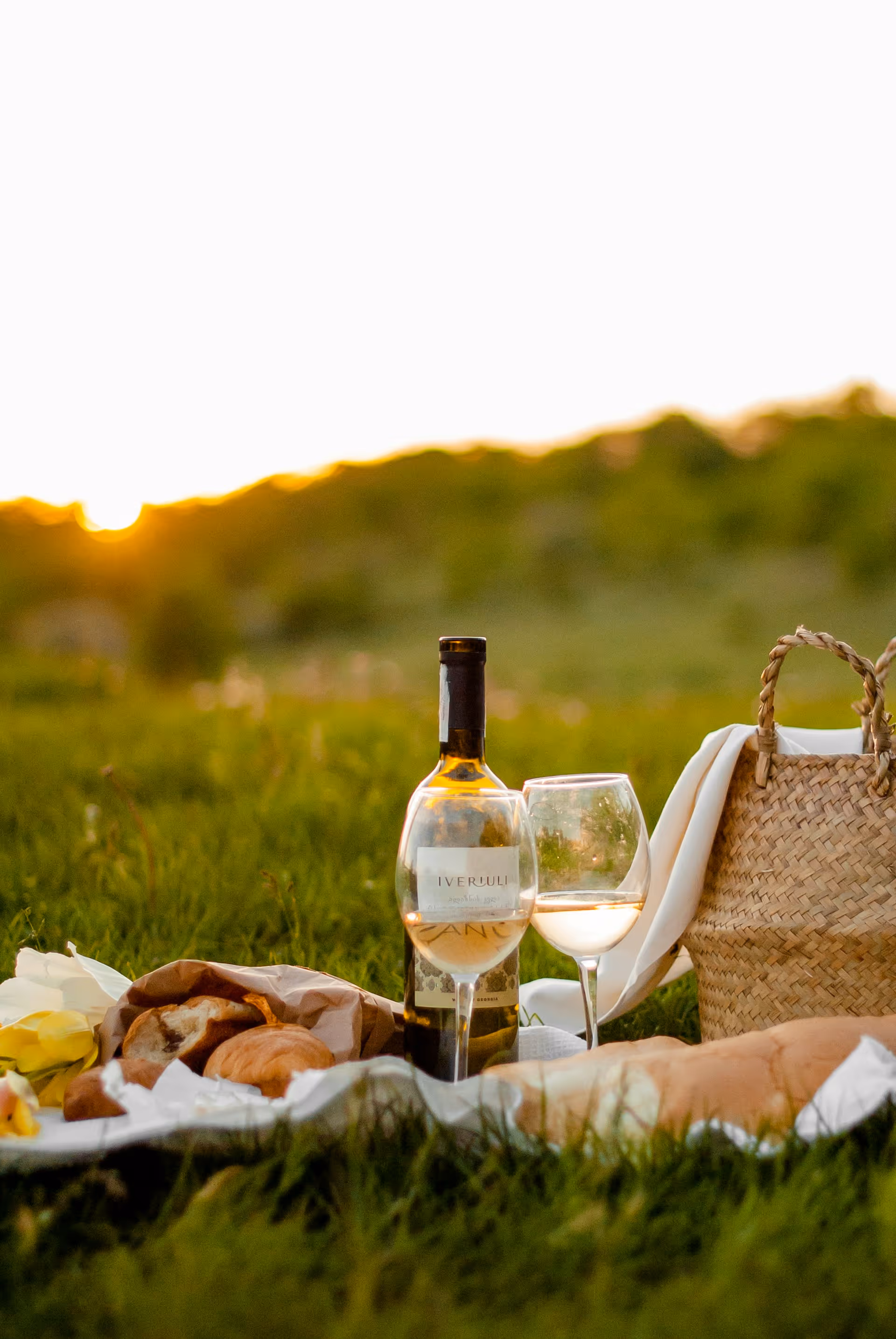 Wine bottle, two glasses of white wine, bread, and a woven basket on a blanket in a grassy field at sunset.