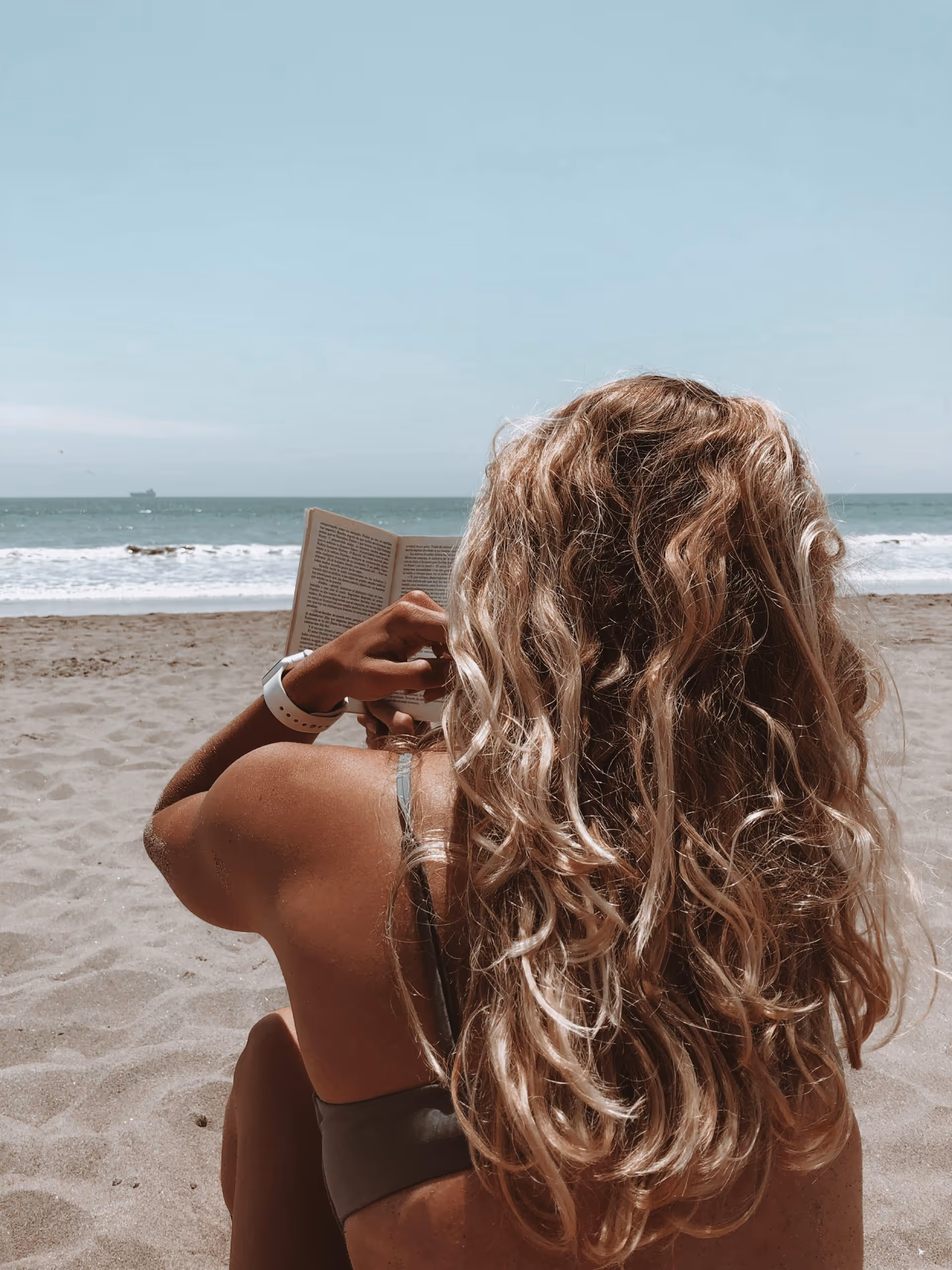 Person with long curly hair reading a book while sitting on a sandy beach facing the ocean.