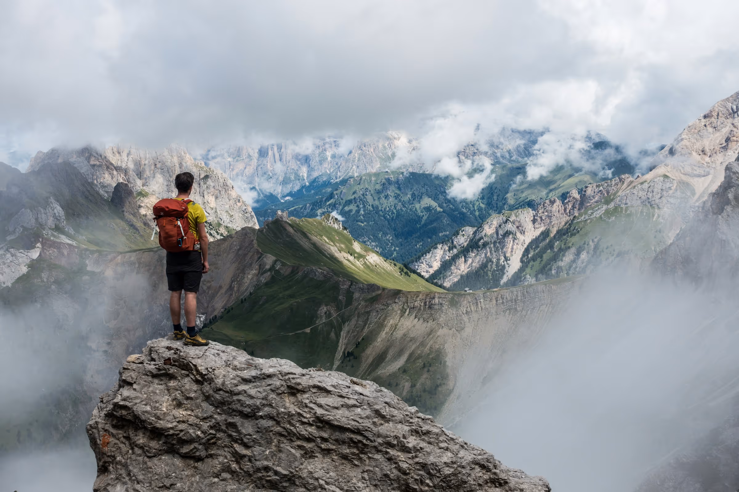 Hiker with red backpack standing on a rocky ledge overlooking fog-covered mountains and green valleys under a cloudy sky.