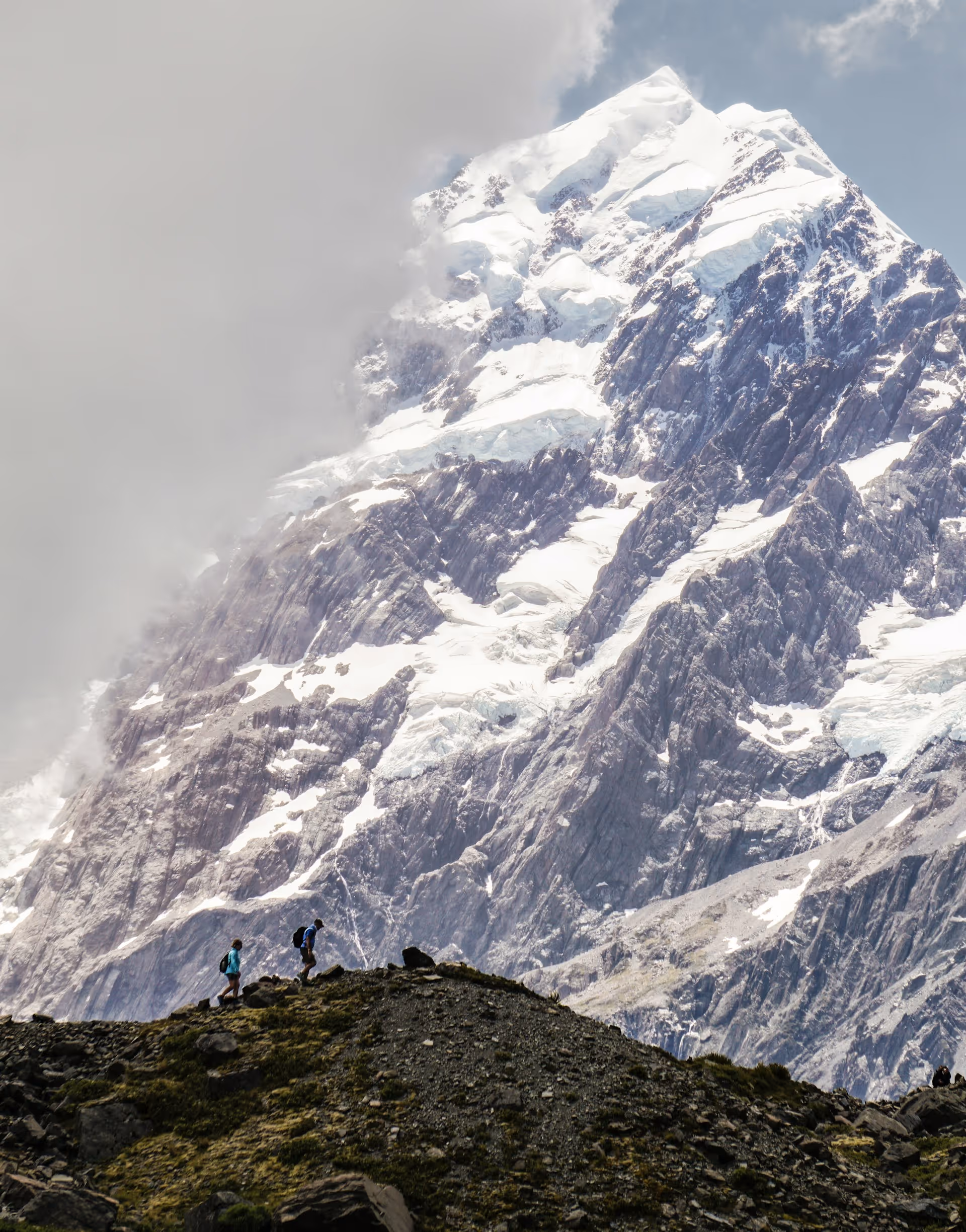 Two hikers walking along a rocky ridge with a towering snow-covered mountain peak and clouds in the background.