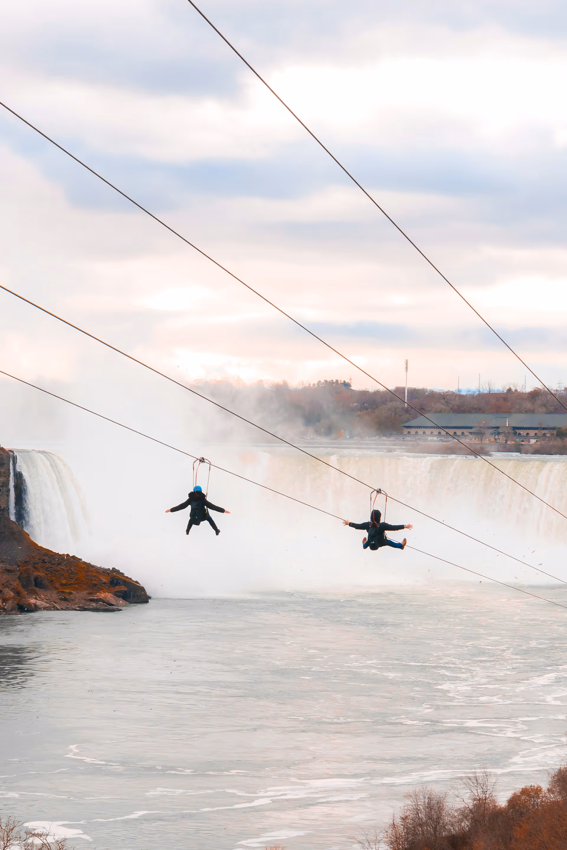Two individuals ziplining over a river with a large waterfall and mist in the background under a cloudy sky.