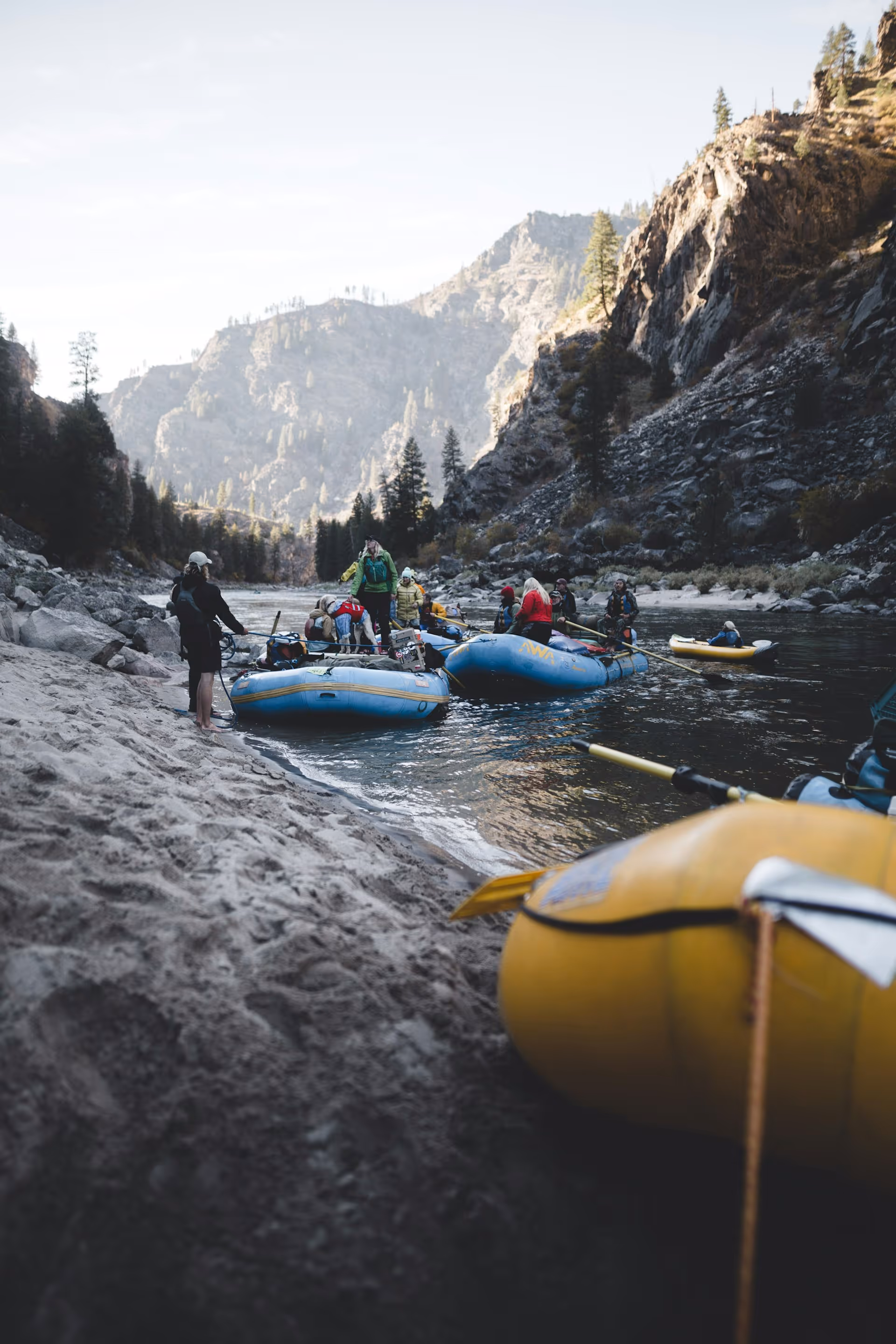 Group of people preparing blue and yellow rafts on a riverbank surrounded by rocky mountains and pine trees.