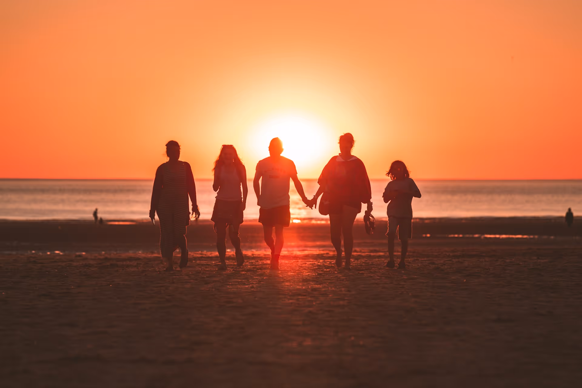 Silhouettes of five people walking hand in hand on a beach at sunset with the sun low on the horizon.