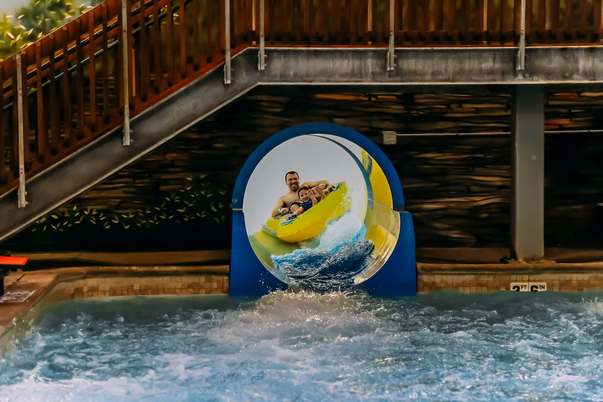 Man and child riding a yellow inflatable raft exiting a water slide into a pool.