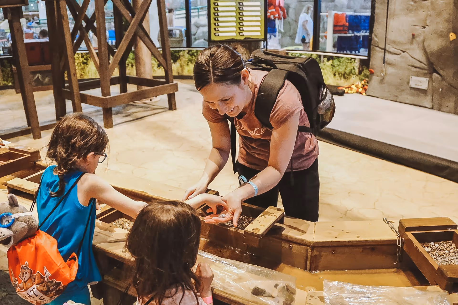 Woman smiling and showing small objects to two children playing at an indoor water and sand activity station.