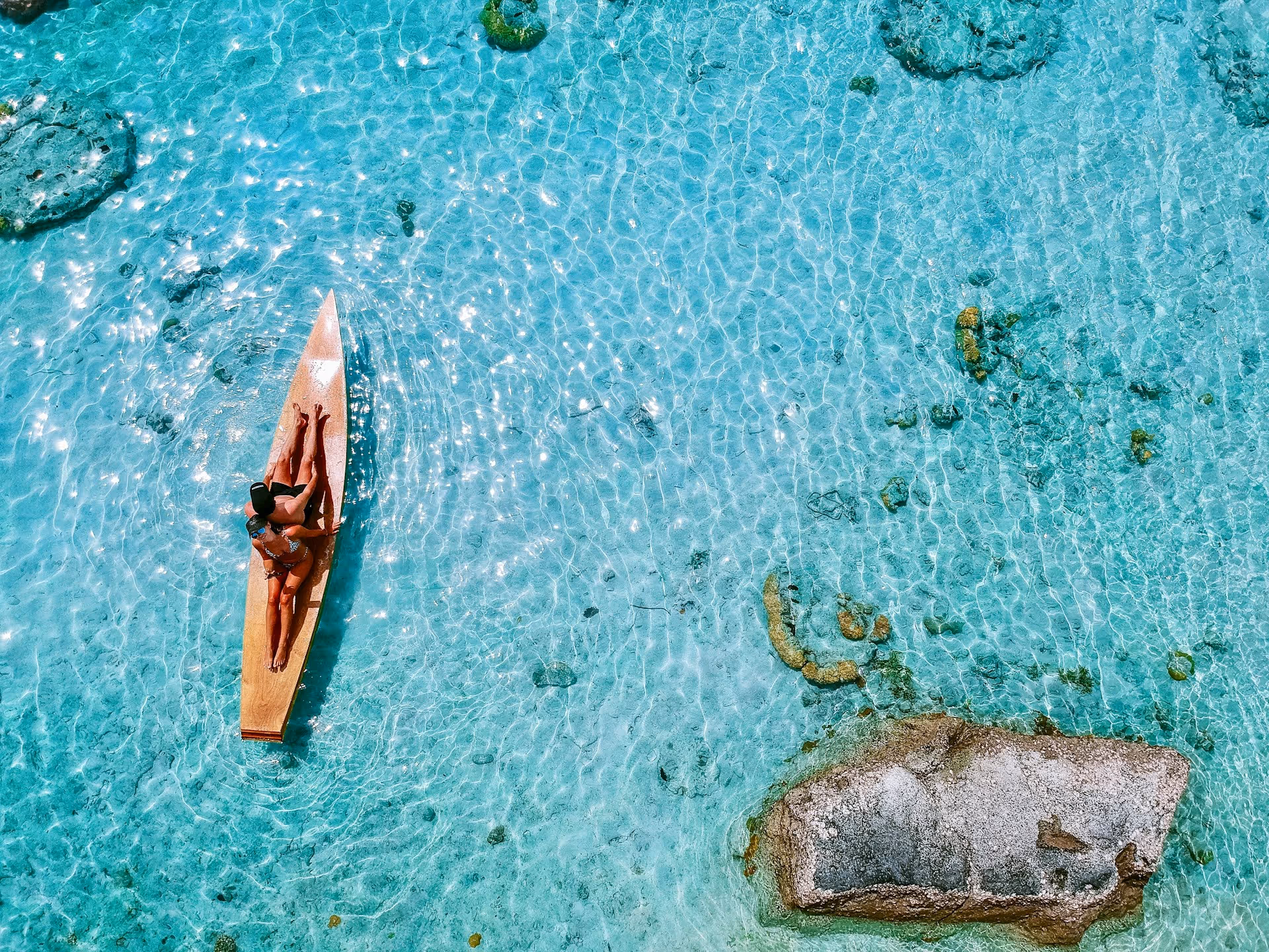 A person relaxing on a wooden paddleboard in clear turquoise water with visible rocks beneath.