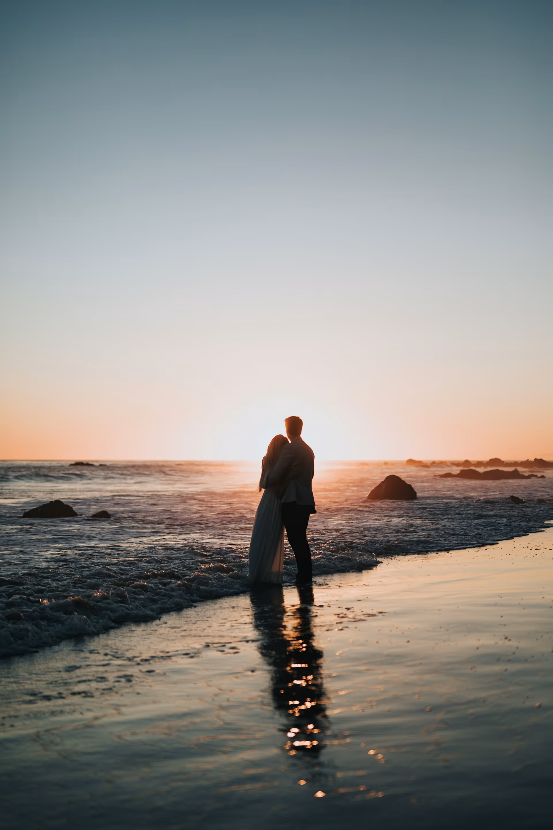 Couple embracing and standing in shallow ocean water at sunset with reflections on wet sand.