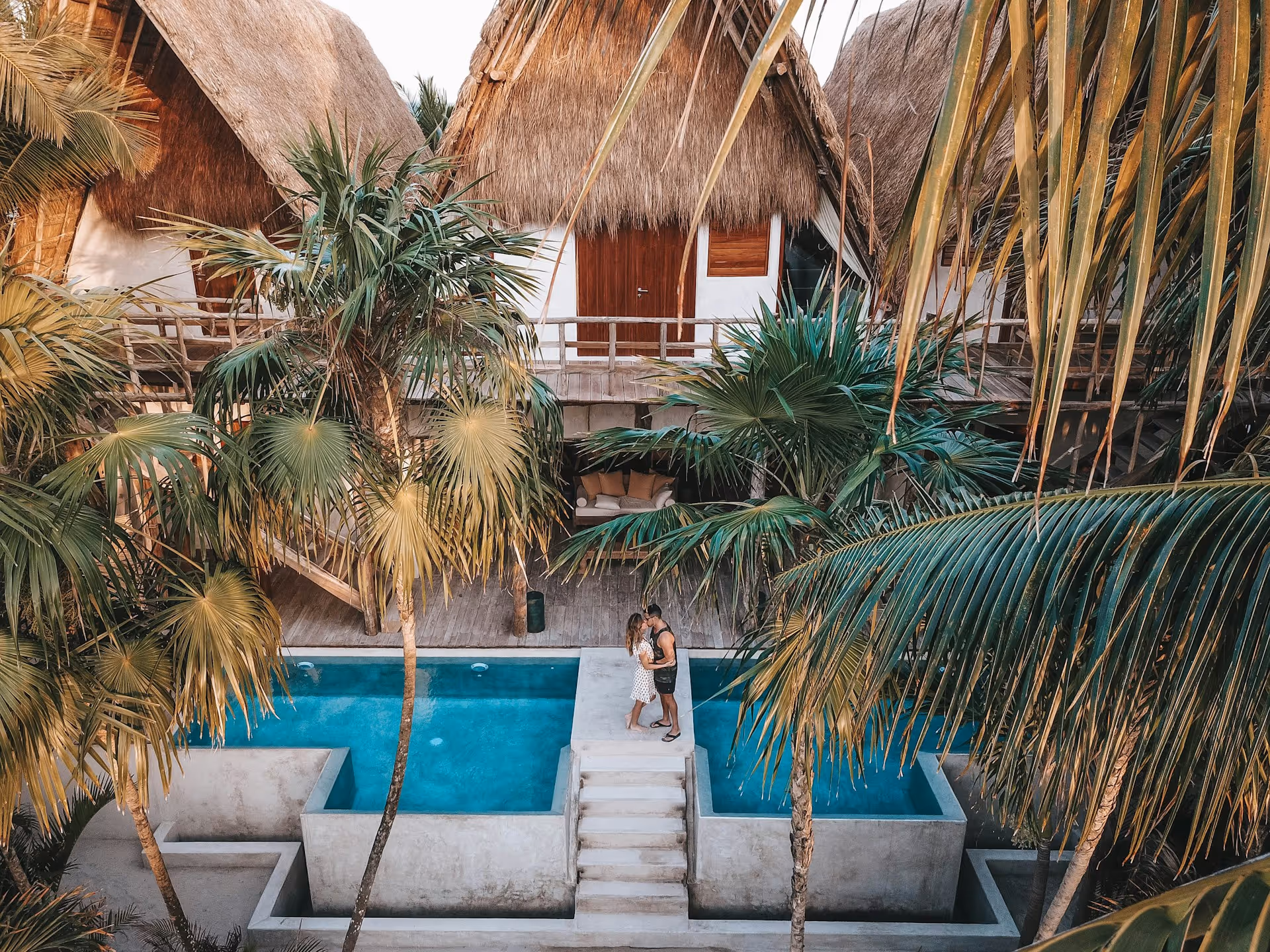 Couple standing on a walkway between two blue rectangular pools surrounded by palm trees and tropical thatched-roof buildings.