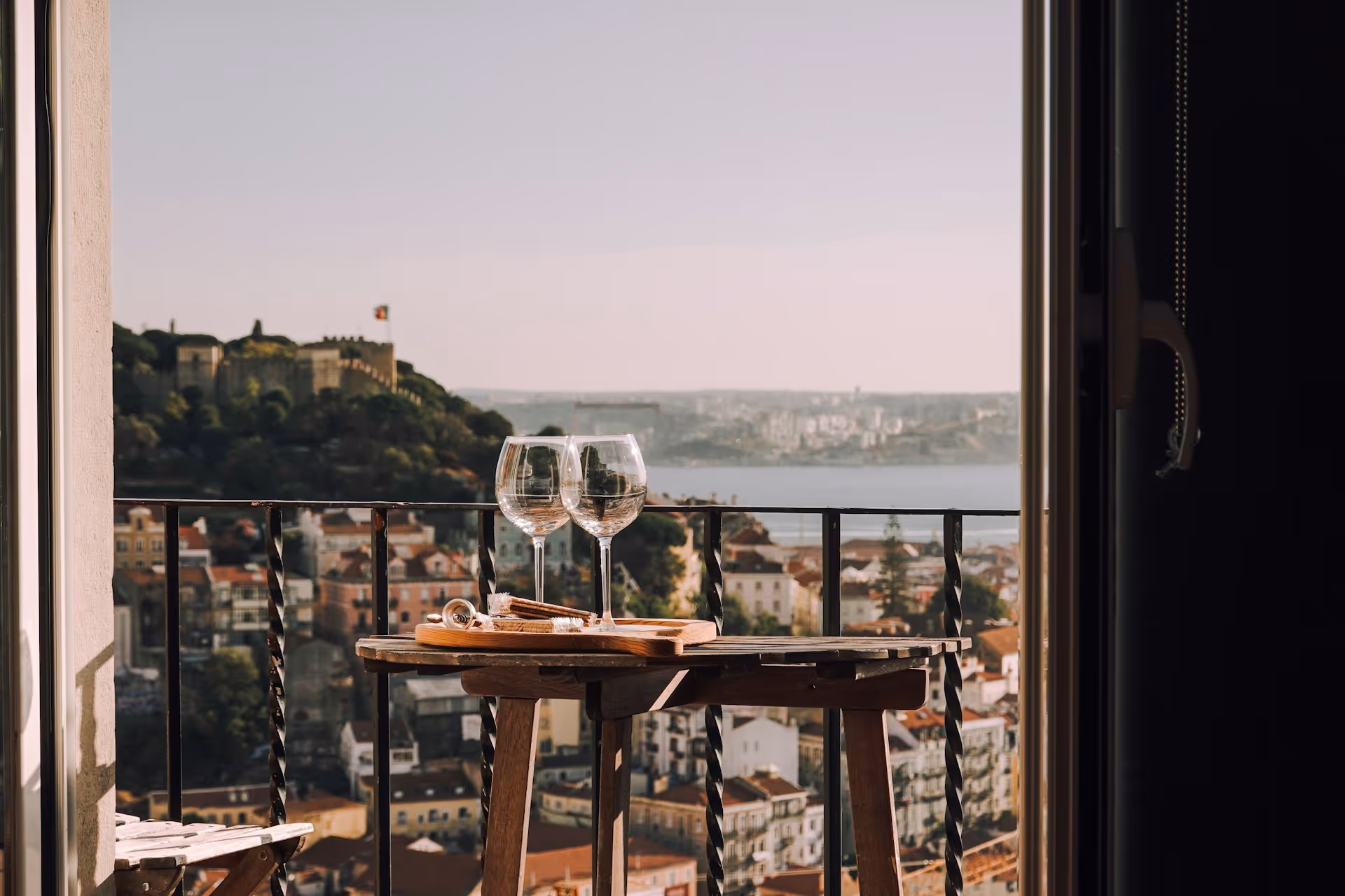 Two empty wine glasses and a small serving tray with snacks on a wooden table on a balcony overlooking a city with a castle on a hill and a river in the background.