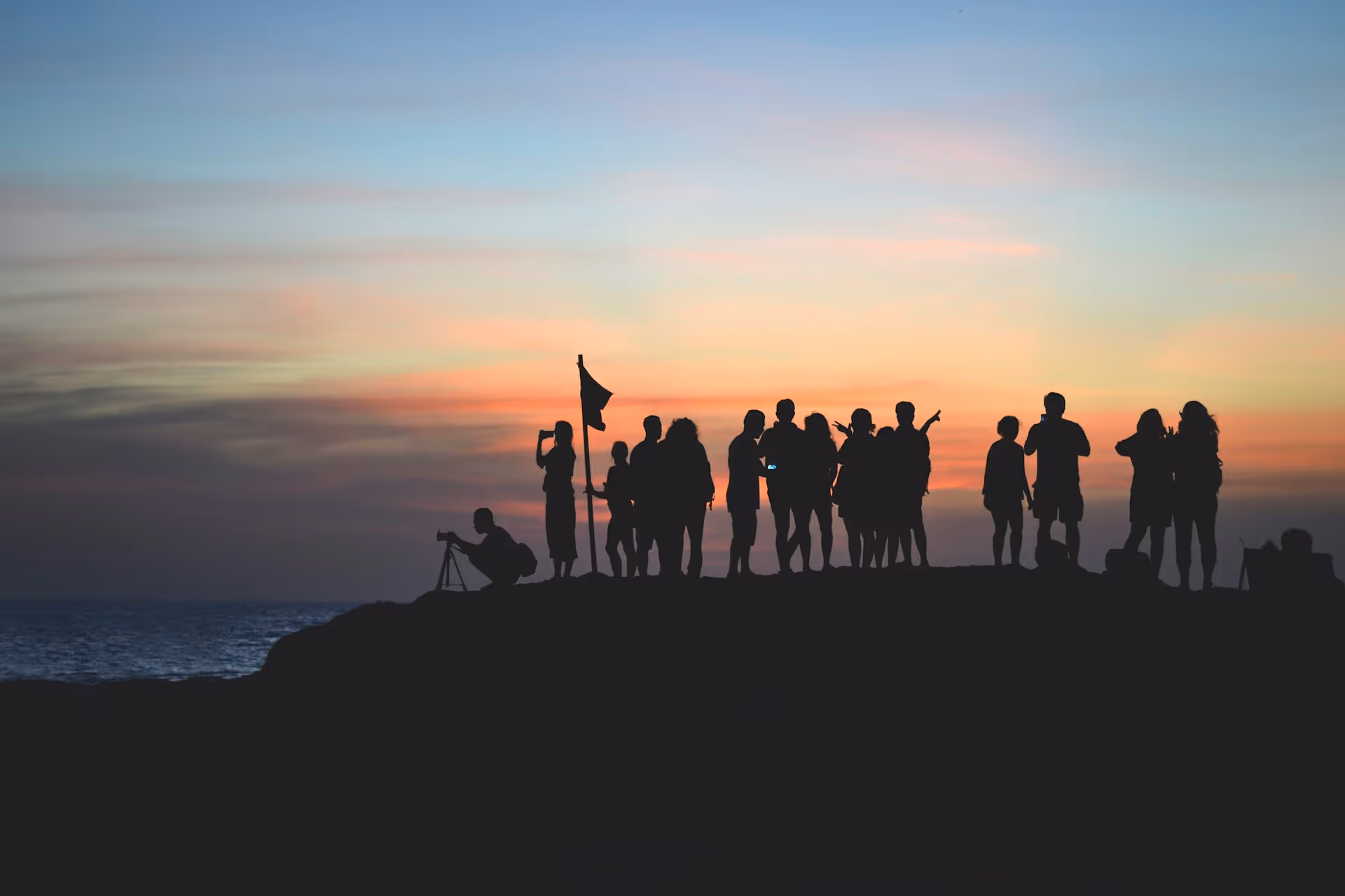 Group of people silhouetted on a hill at sunset near the ocean, some taking photos and holding a flag.