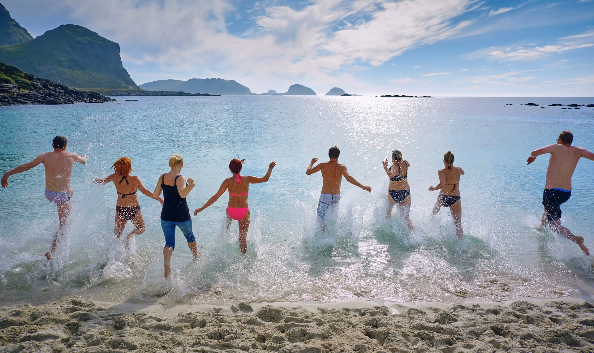 Seven people jumping into clear ocean water from a sandy beach with rocky hills and islands in the background under a partly cloudy sky.