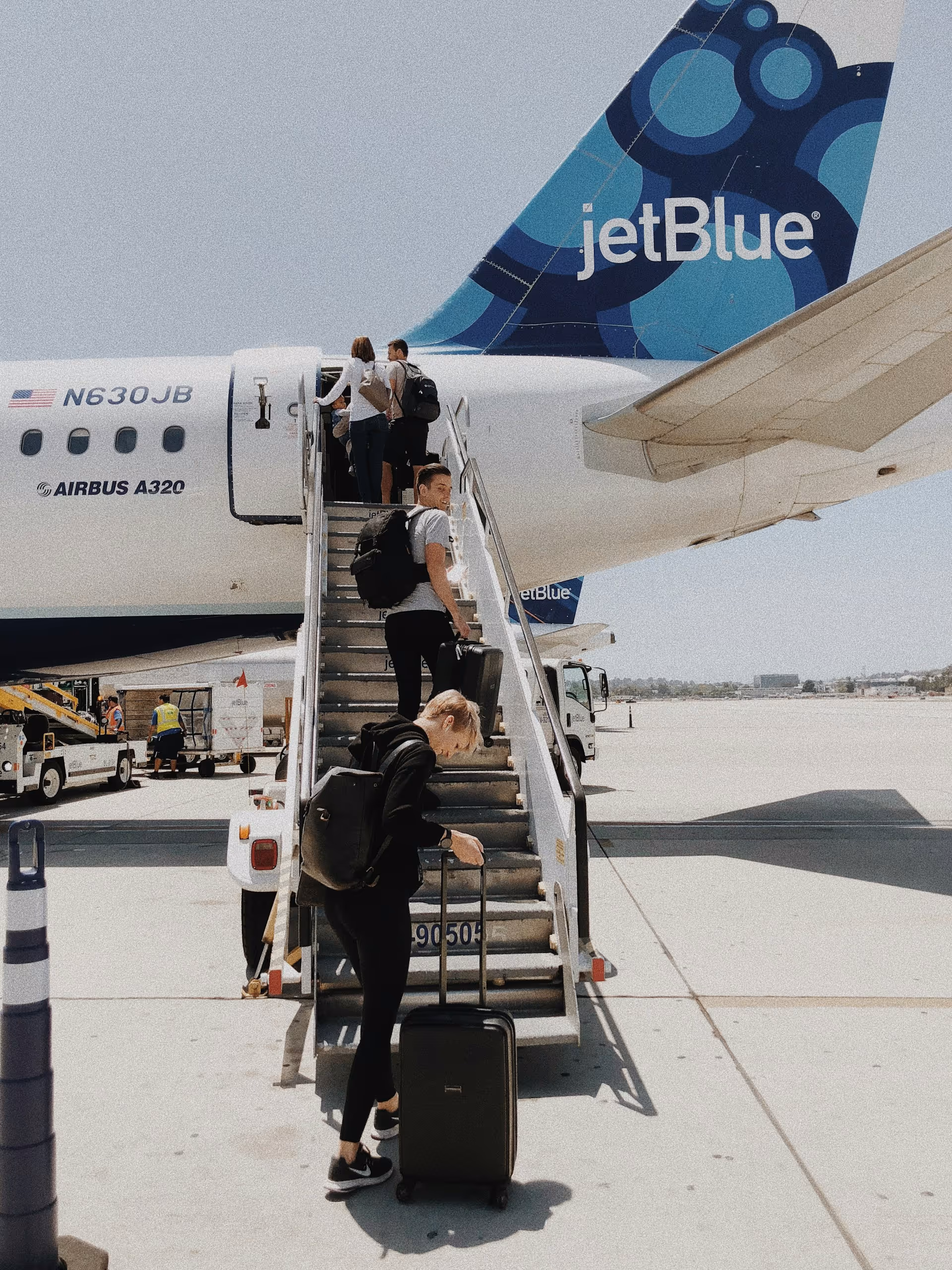 Passengers with luggage boarding a JetBlue Airbus A320 plane via stairs on an airport tarmac.