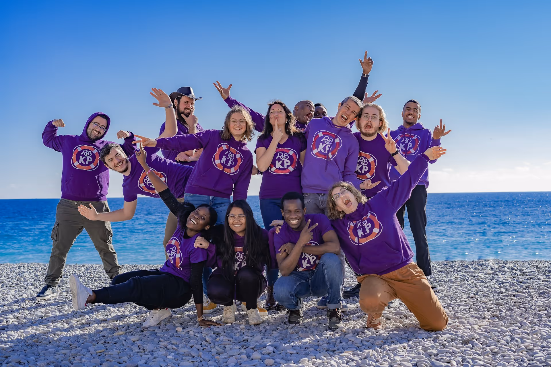 Group of people wearing matching purple shirts and hoodies posing playfully on a rocky beach with the ocean and clear blue sky in the background.