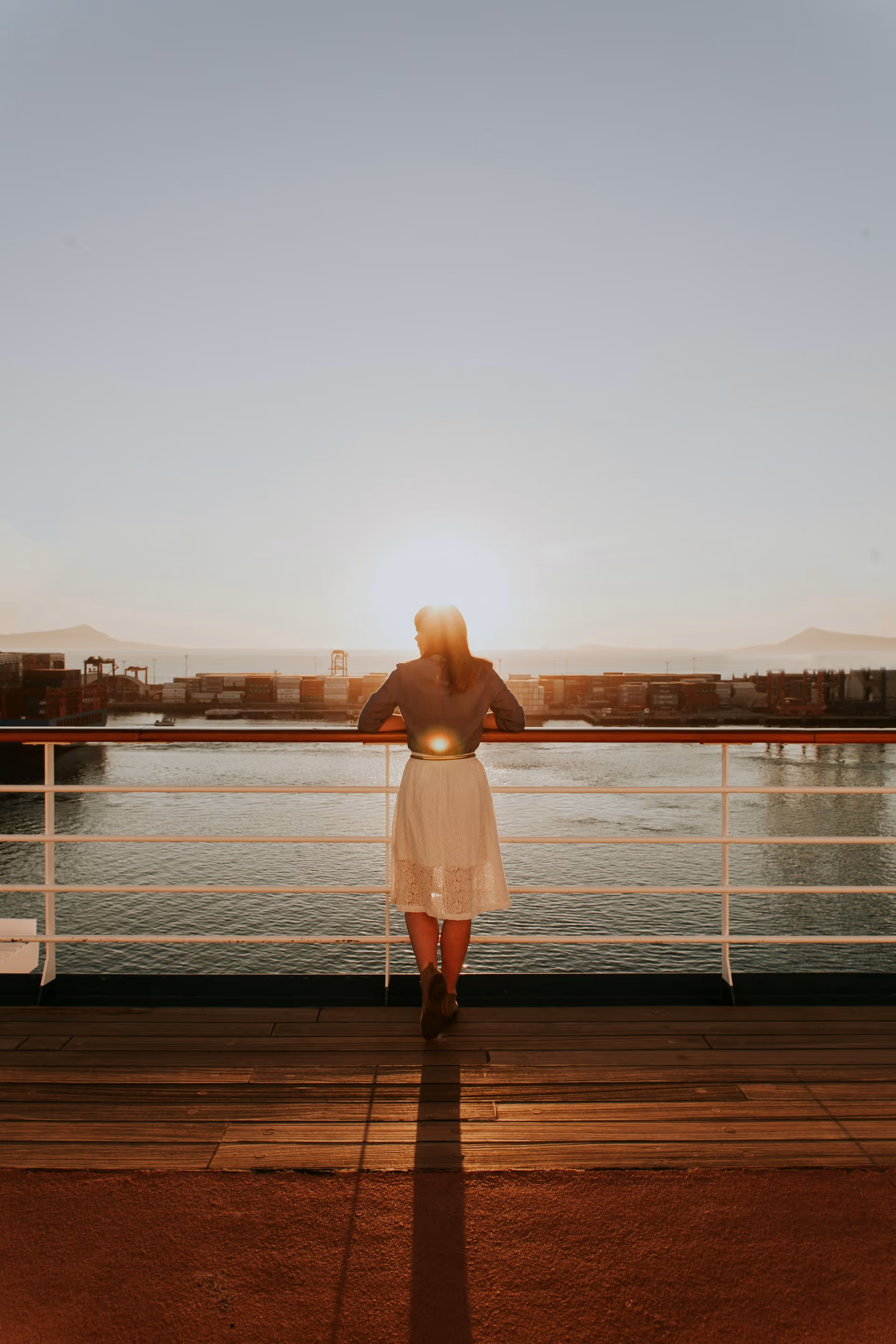 Woman in a white skirt and jacket leaning on a railing, watching the sunset over a harbor with shipping containers.