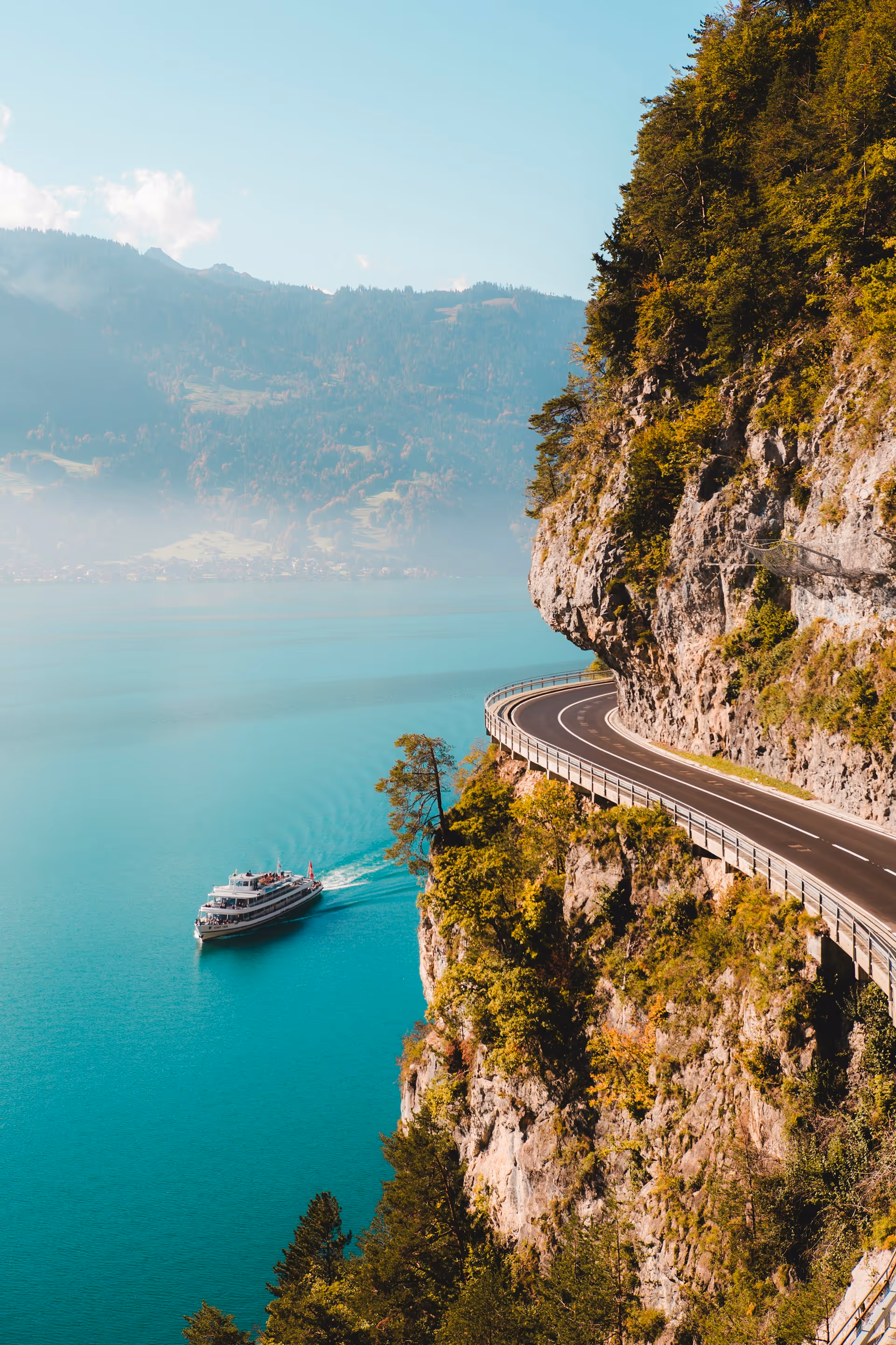 Boat cruising on turquoise lake beside a winding road along a steep, forested cliff.