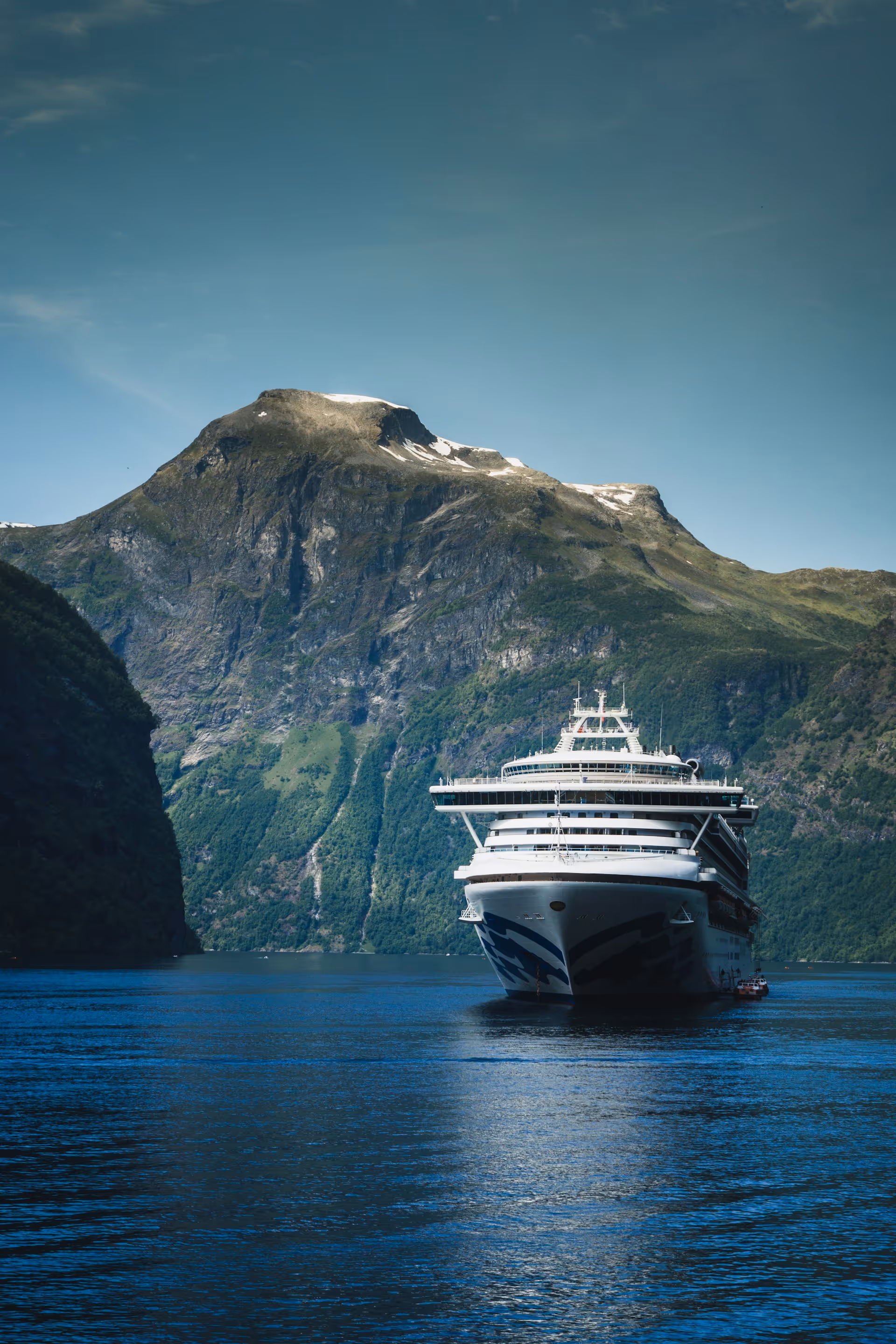 A large white cruise ship sailing on a blue fjord with steep green mountains and a clear sky in the background.