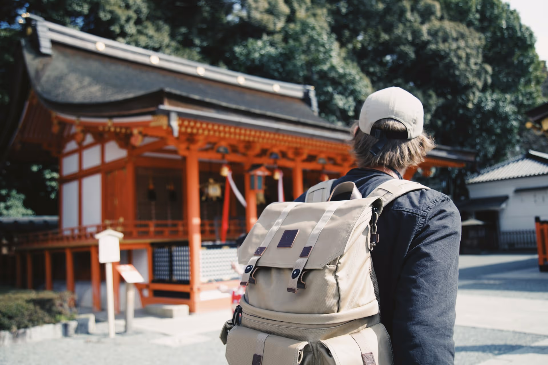 Person wearing a beige backpack and cap standing outdoors near a traditional Japanese shrine with orange wooden pillars.