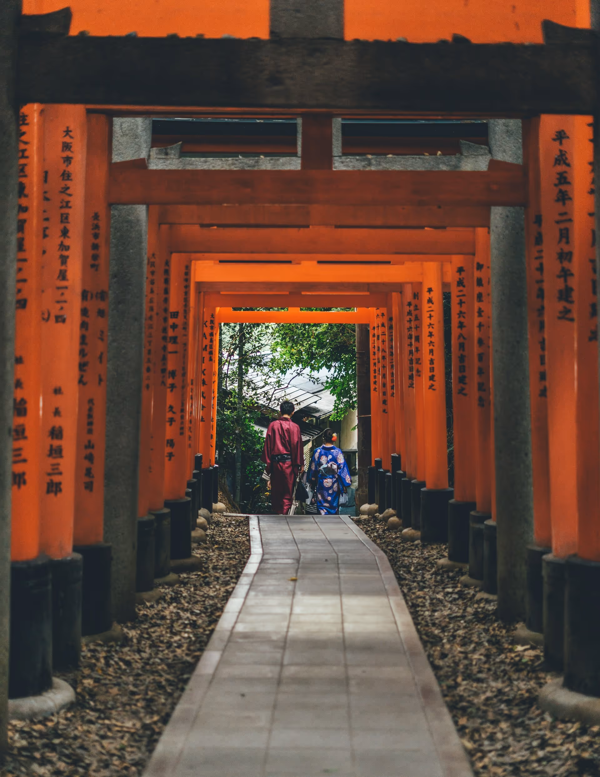 Two people in traditional Japanese attire walking under a series of vibrant red torii gates on a stone path.