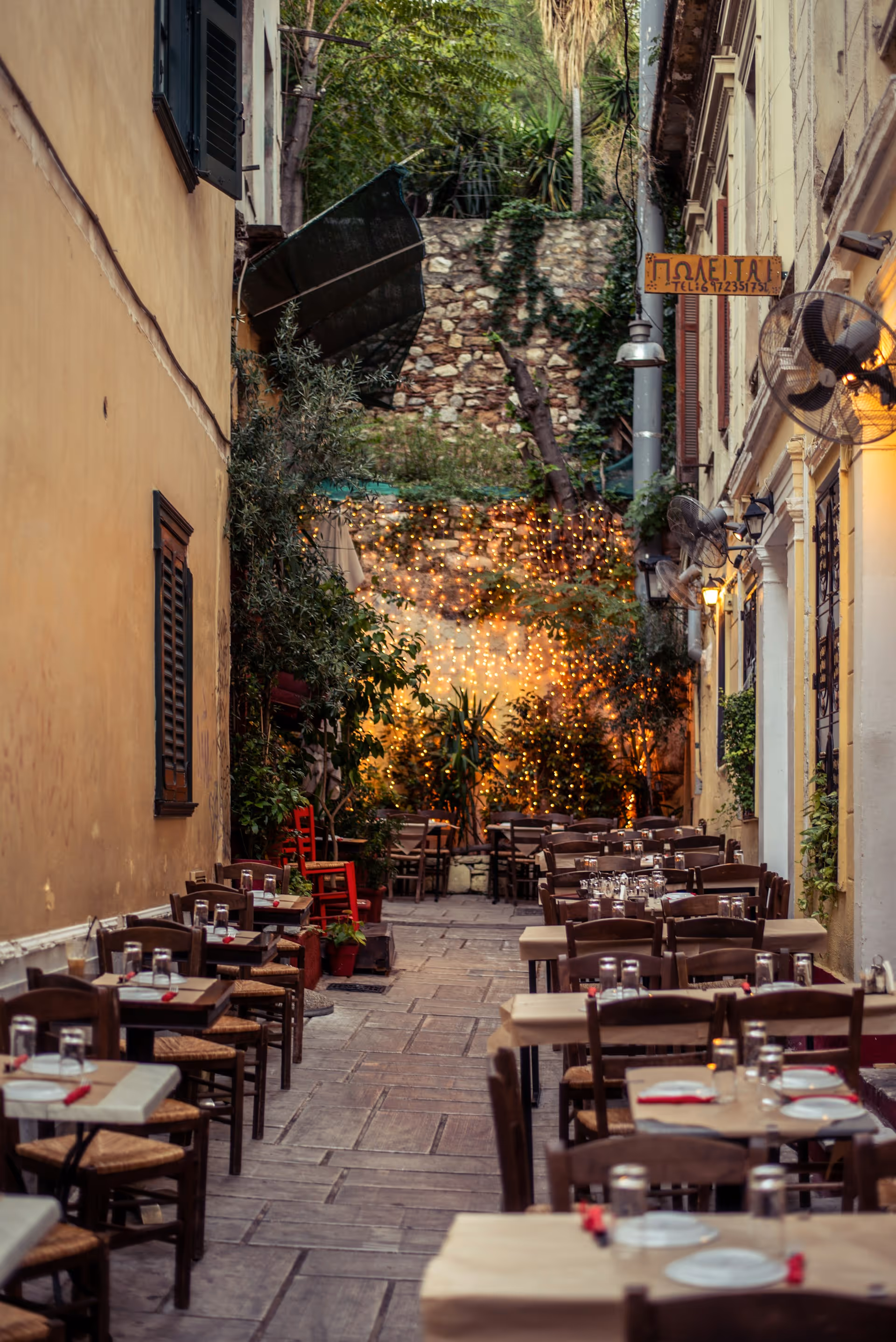 Outdoor street cafe with neatly arranged wooden tables and chairs, string lights hanging on a stone wall in the background.