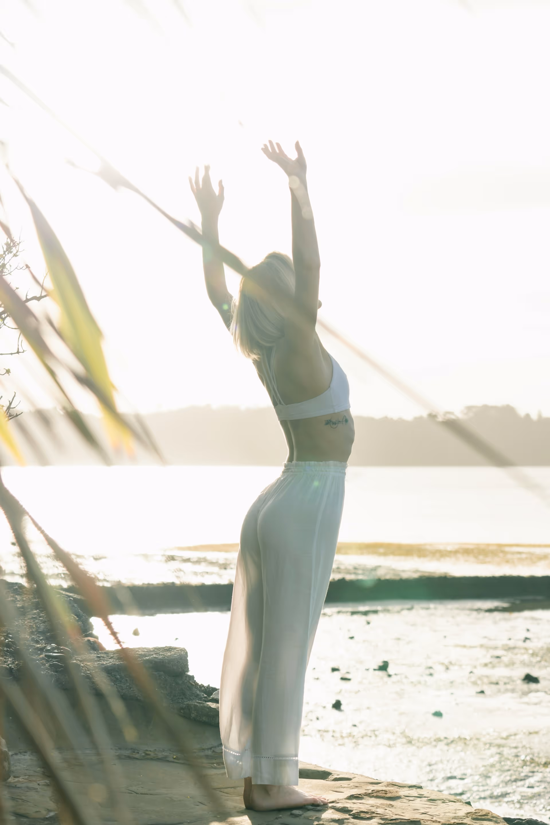 Woman in white pants and sports bra stretching with arms raised by a sunlit lakeside.