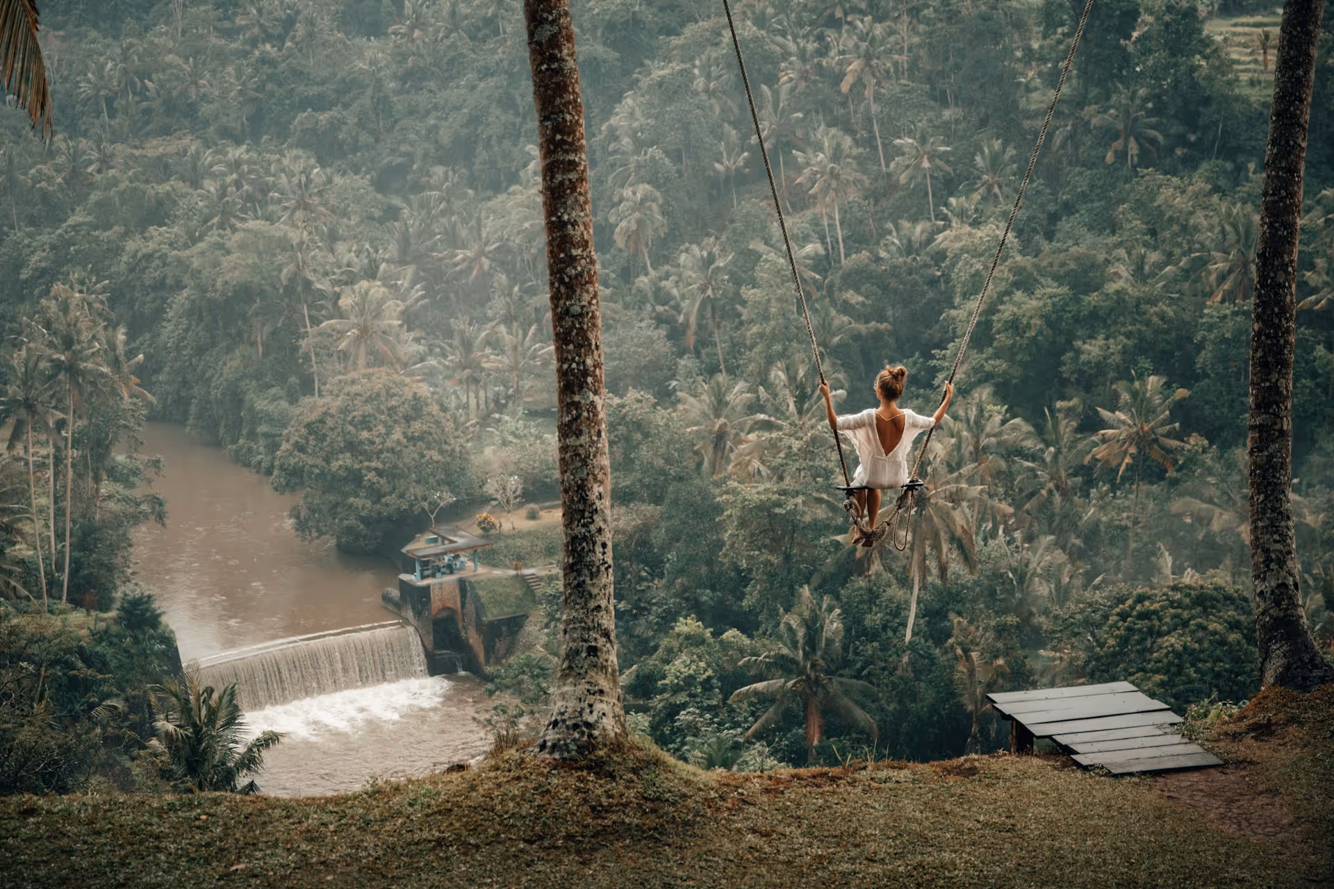 Woman in white dress swinging on a large swing overlooking a lush tropical forest and river with a small waterfall.