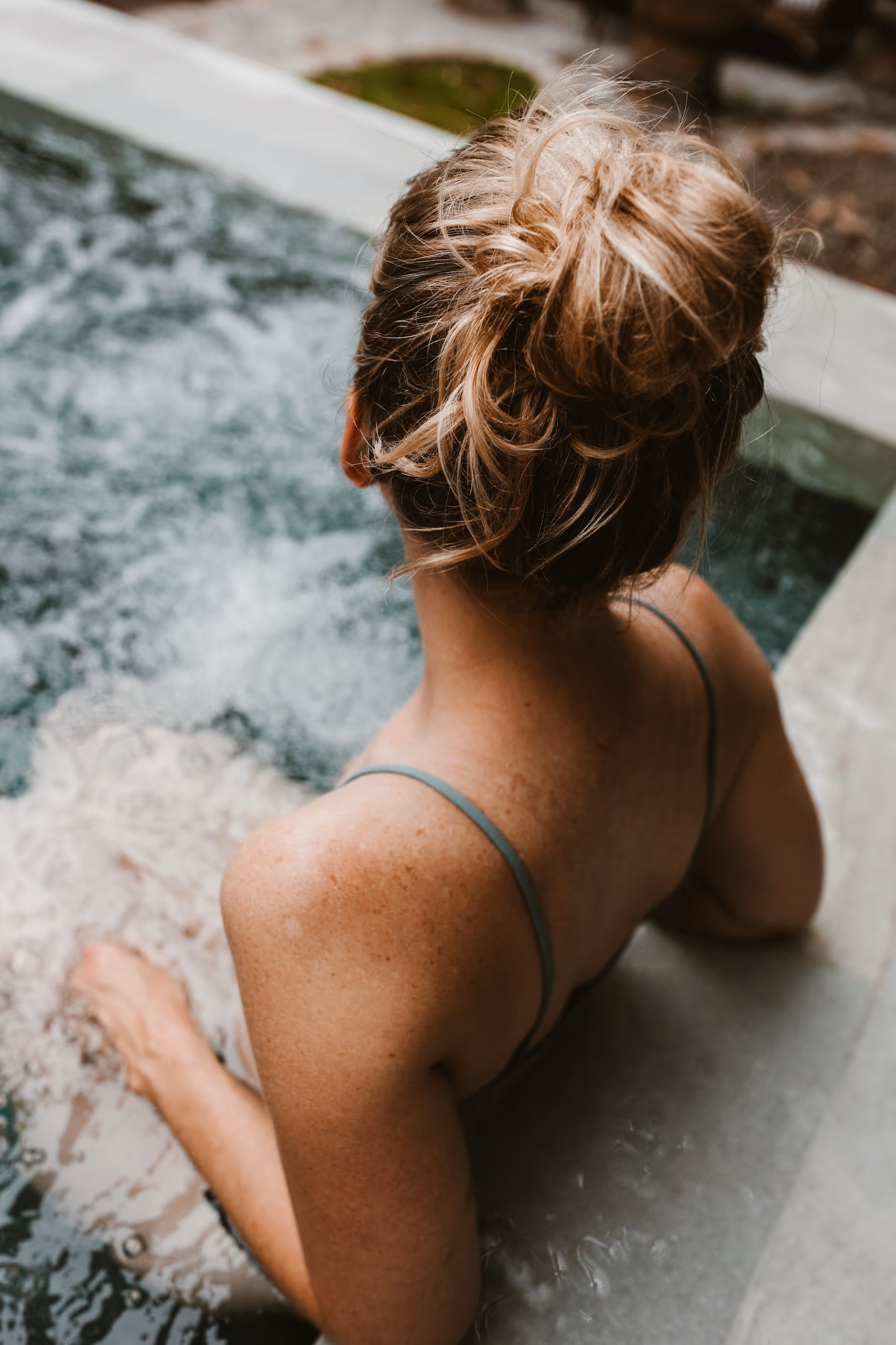 Woman with blonde hair in a bun relaxing in a bubbling hot tub, viewed from behind.