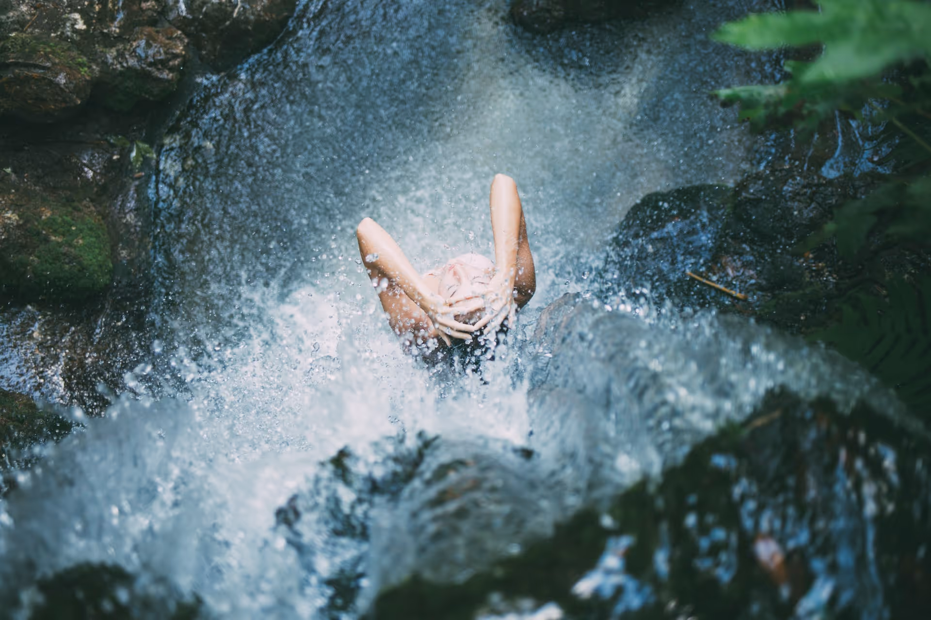 Person standing under a waterfall with hands on their head, surrounded by splashing water and rocks.