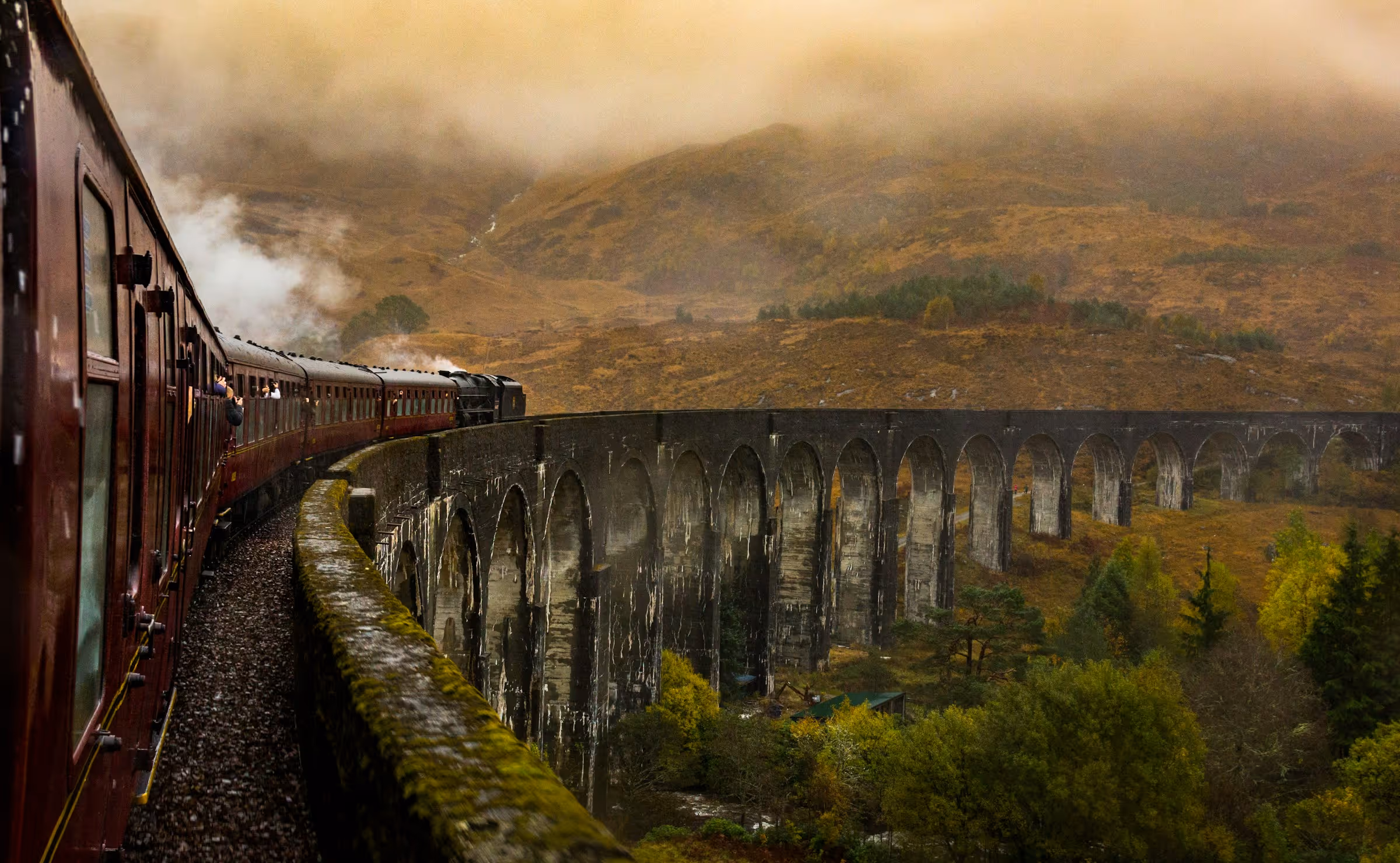 Vintage steam train crossing a curved stone viaduct over a lush valley with misty hills in the background.