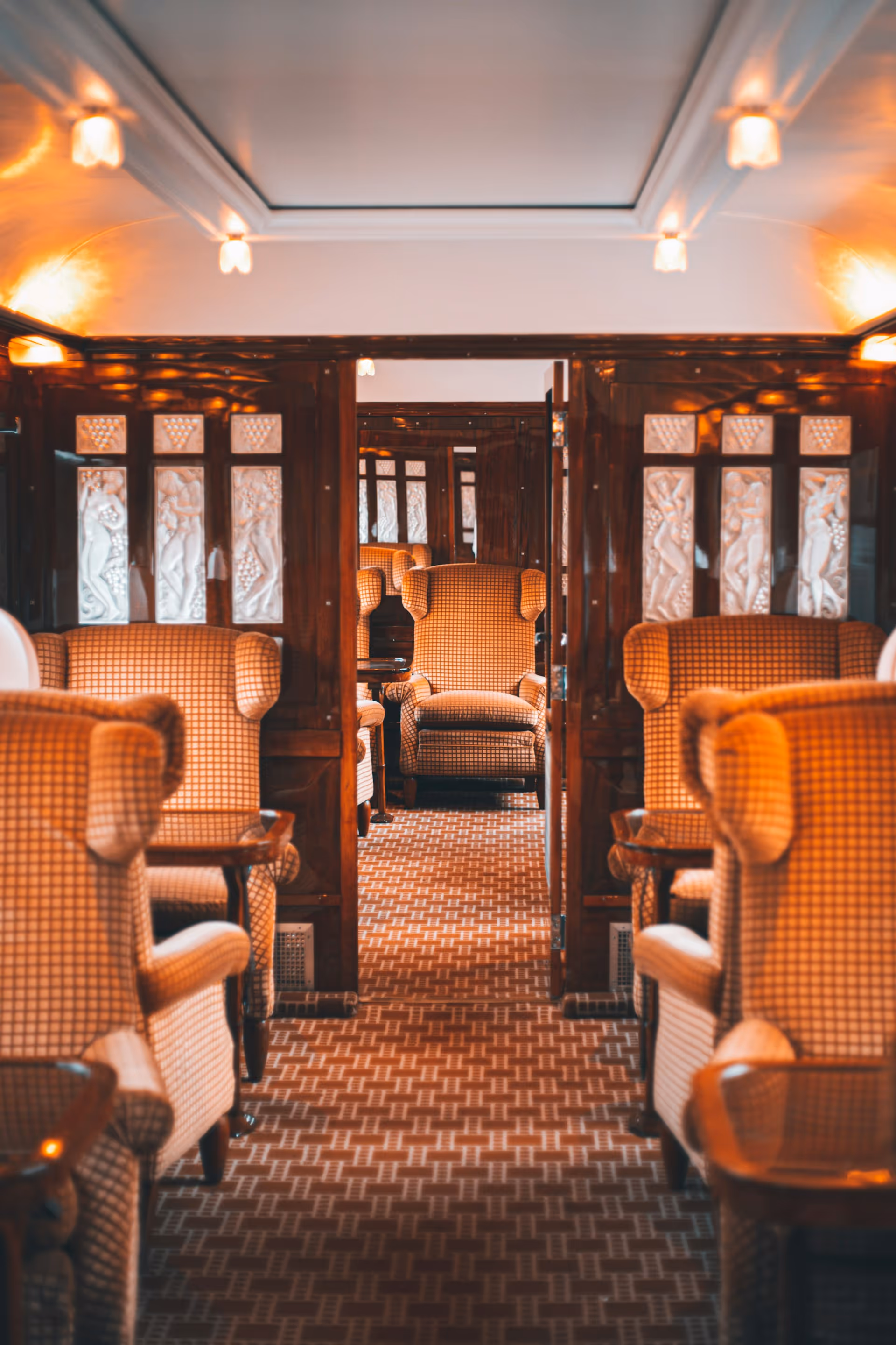 Interior of a vintage train car with patterned armchairs and wooden paneling illuminated by warm lights.