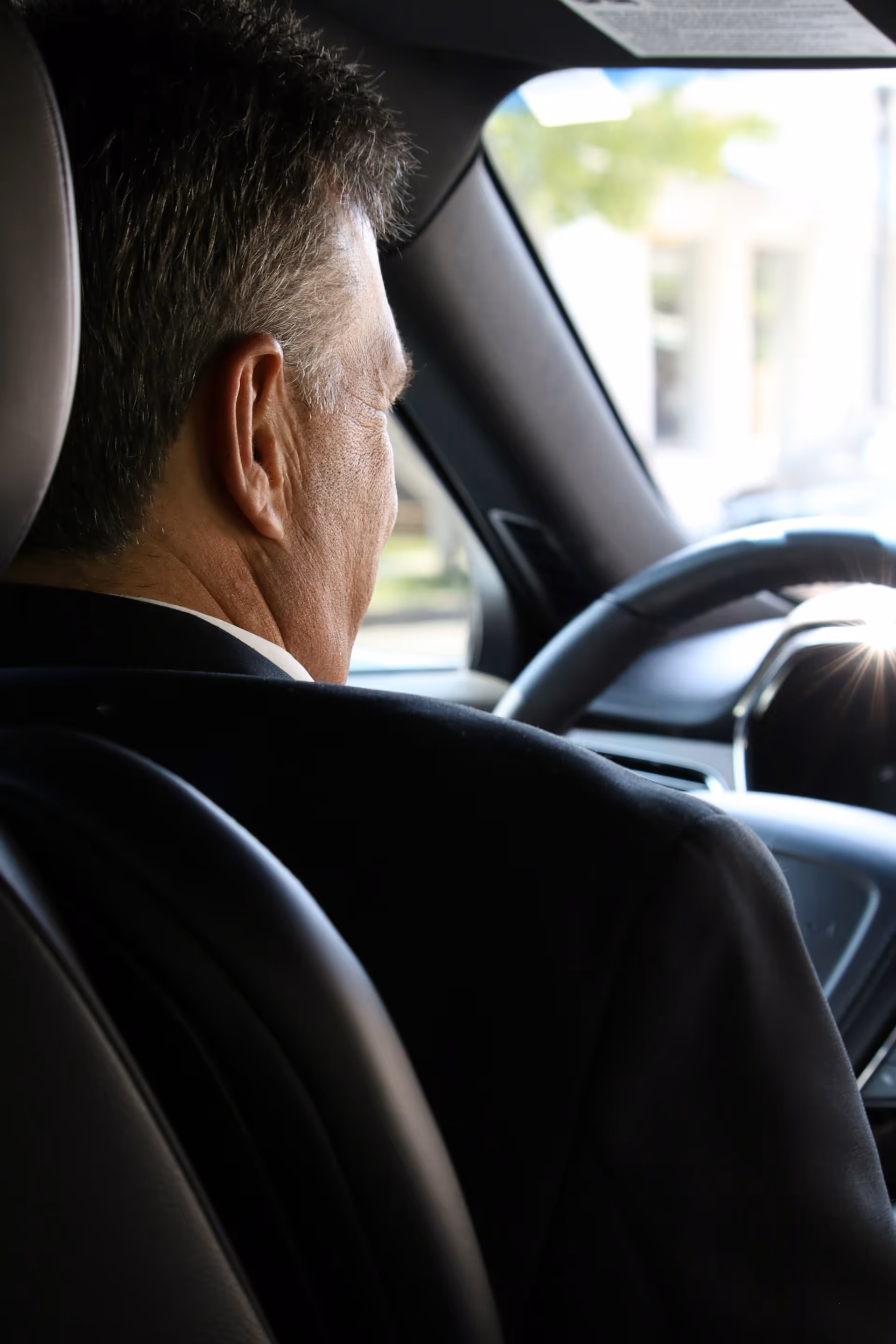Man in a black suit driving a car, viewed from behind his right shoulder.