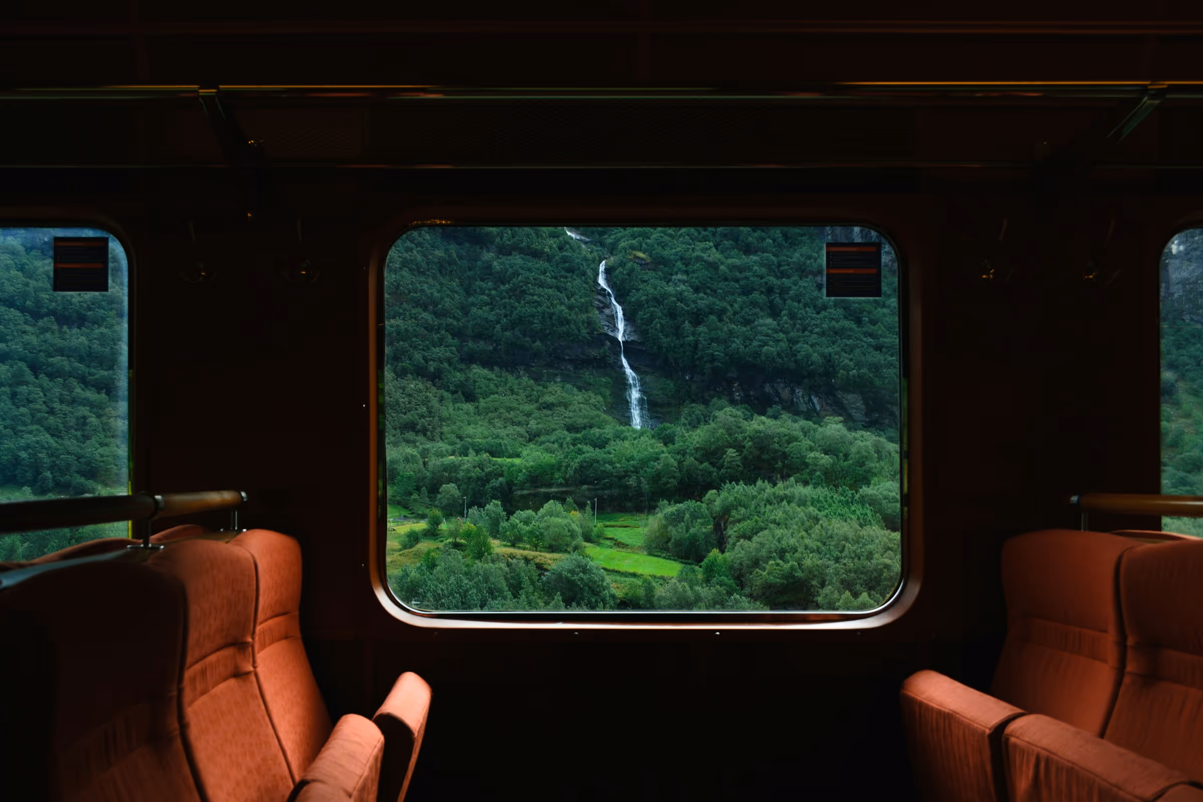 View of a lush green forest and a narrow waterfall through a train window, with empty red seats inside the train.