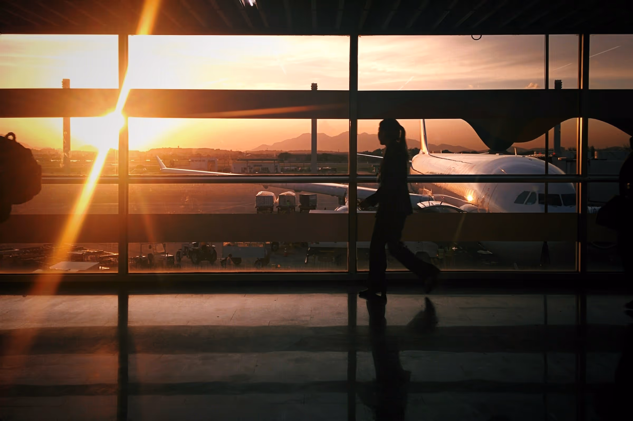 Silhouette of a person walking inside an airport terminal with an airplane and sunset visible outside the window.