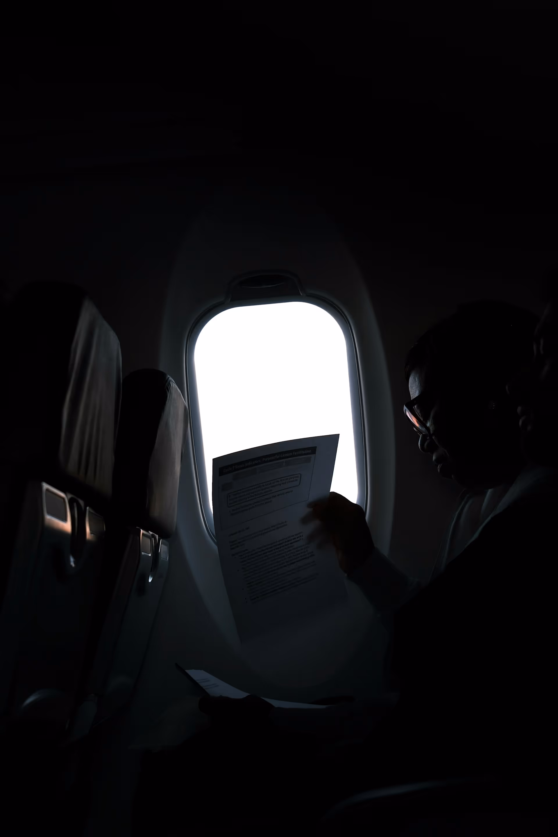 Passenger reading documents beside an airplane window with bright light outside.