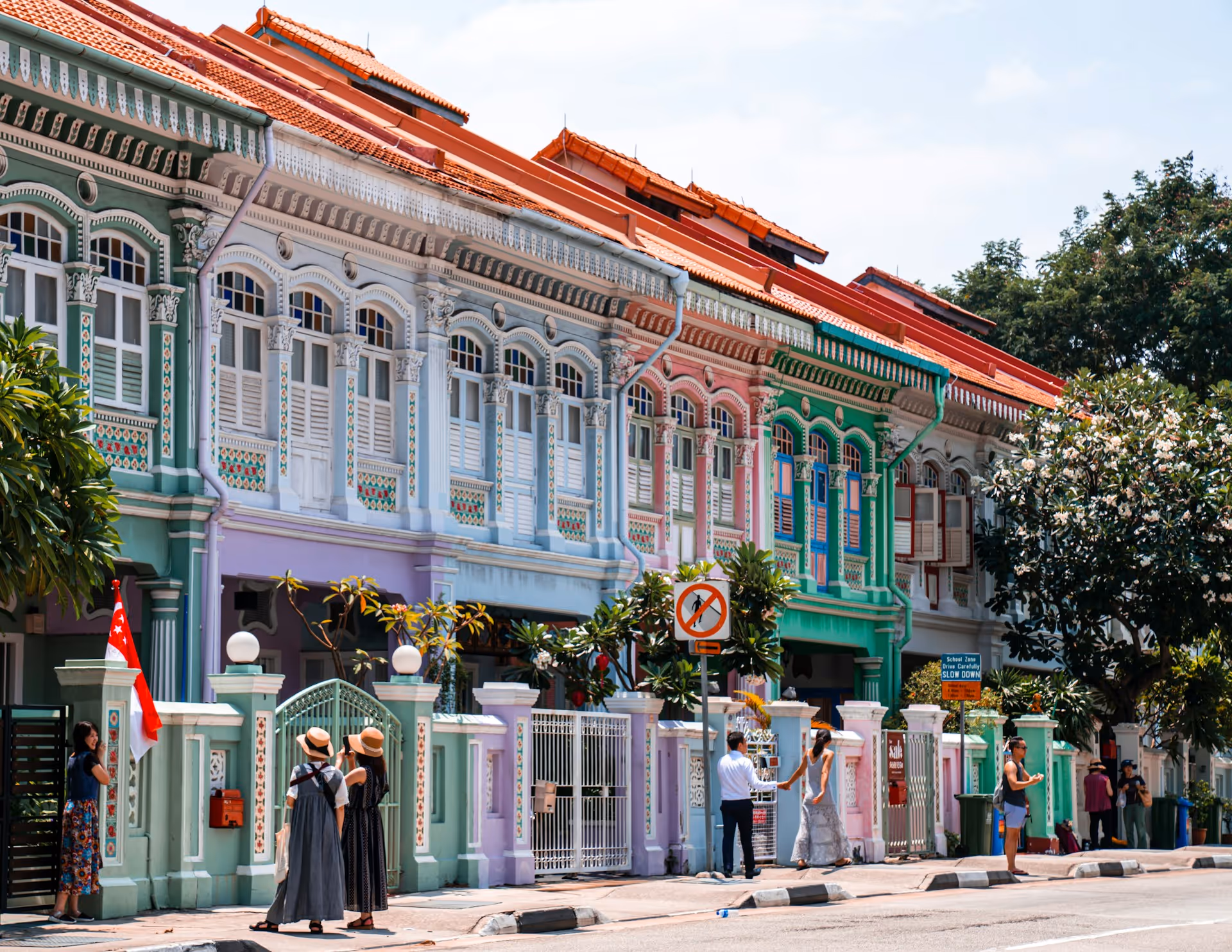 Colorful pastel colonial-style shophouses with ornate details and red tiled roofs along a sunny street with people walking and standing.