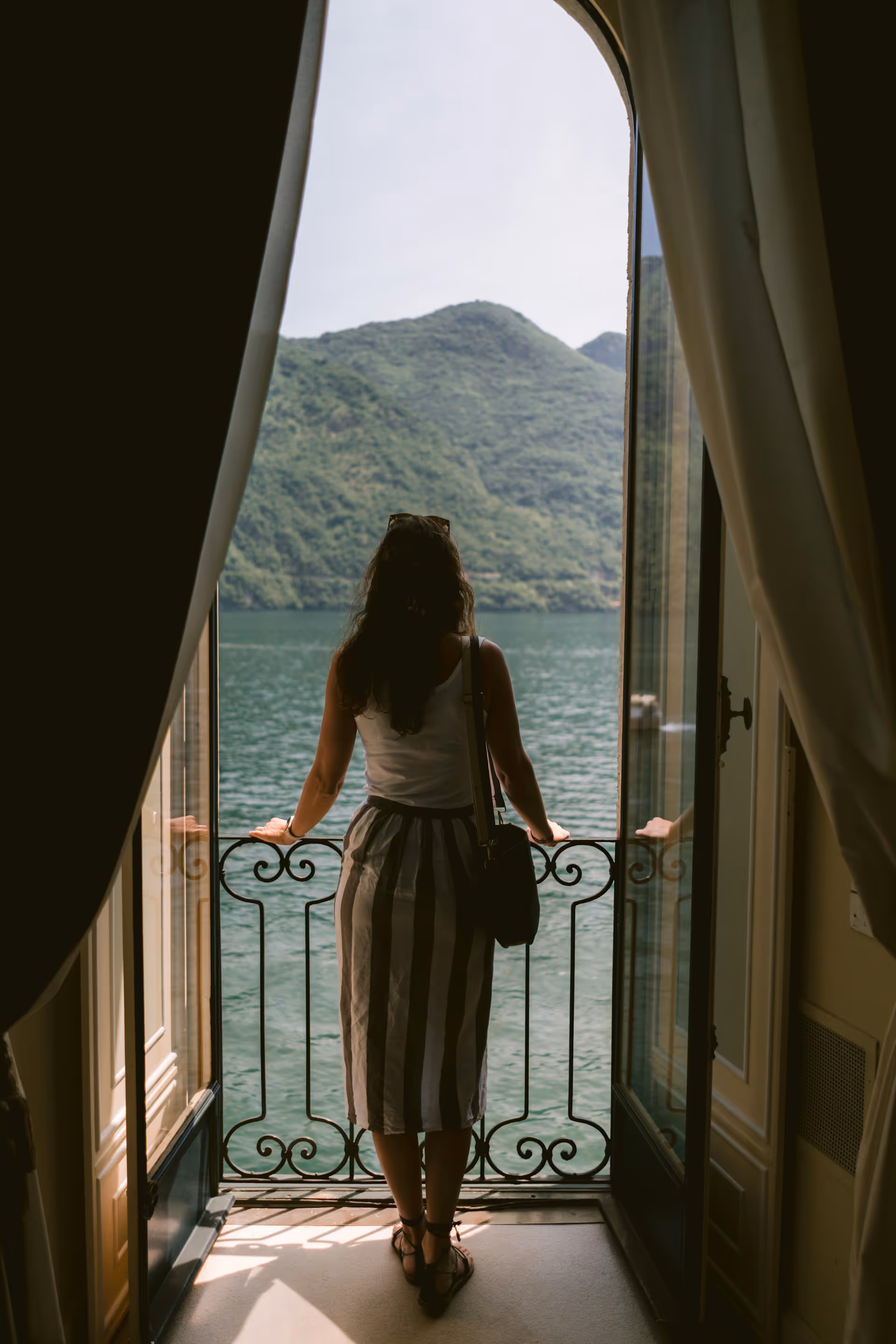 Woman standing on a balcony overlooking a large body of water with green hills in the background.
