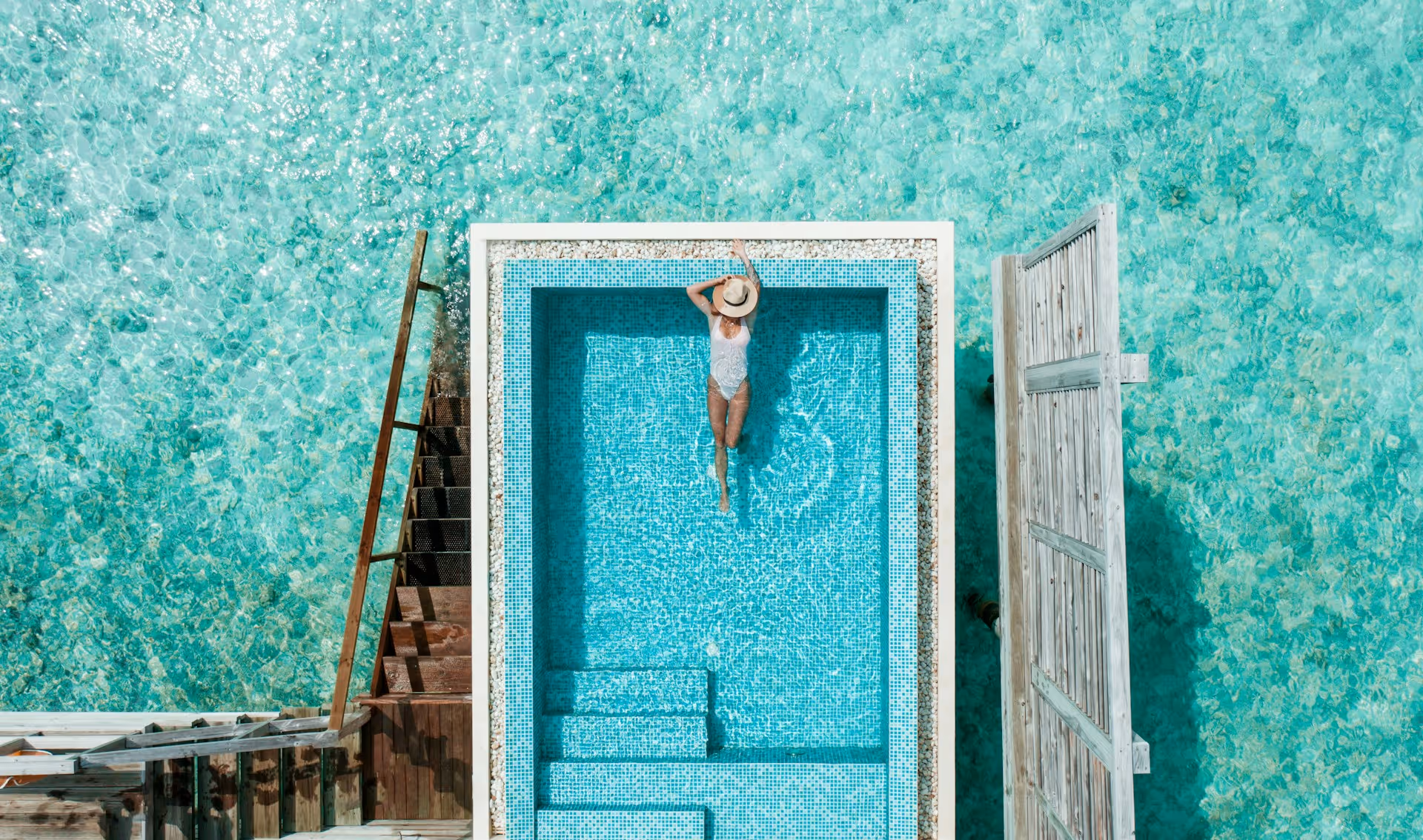 A woman in a white swimsuit and hat relaxing in a small turquoise pool surrounded by clear ocean water.