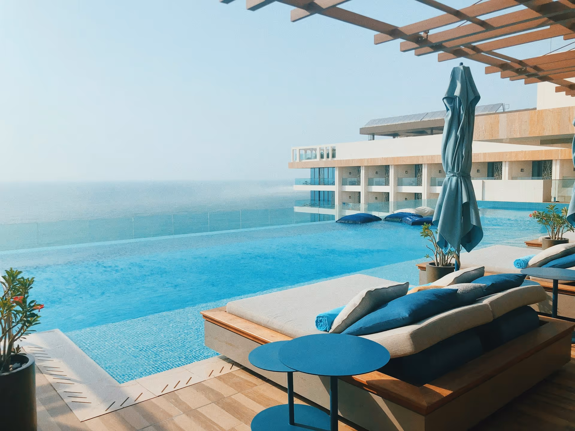 Infinity pool overlooking the ocean with lounge chairs, blue cushions, and closed umbrellas on the deck.