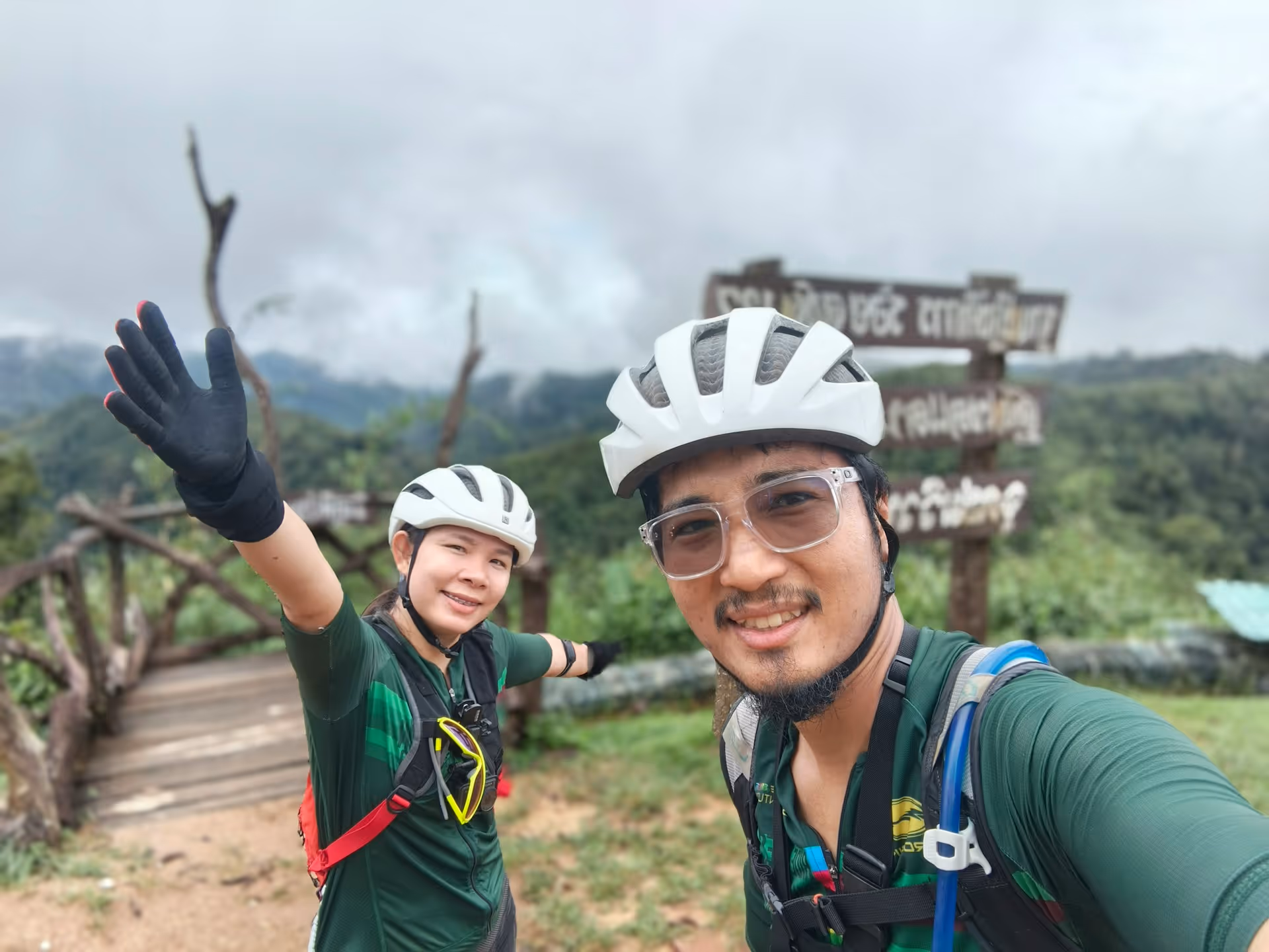 Two cyclists wearing helmets and gear taking a selfie in a green, mountainous outdoor setting with a wooden bridge behind them.