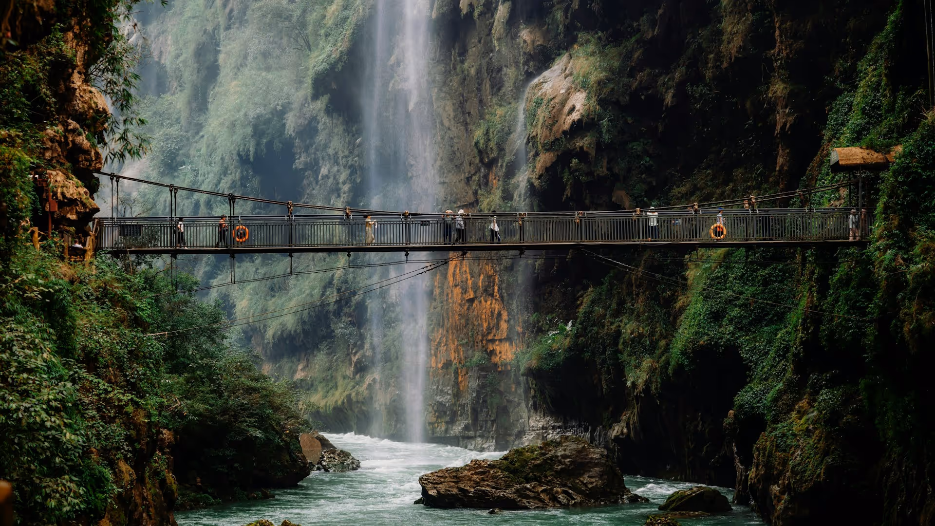 Suspension bridge with people crossing over a river with a waterfall and lush green cliffs in the background.