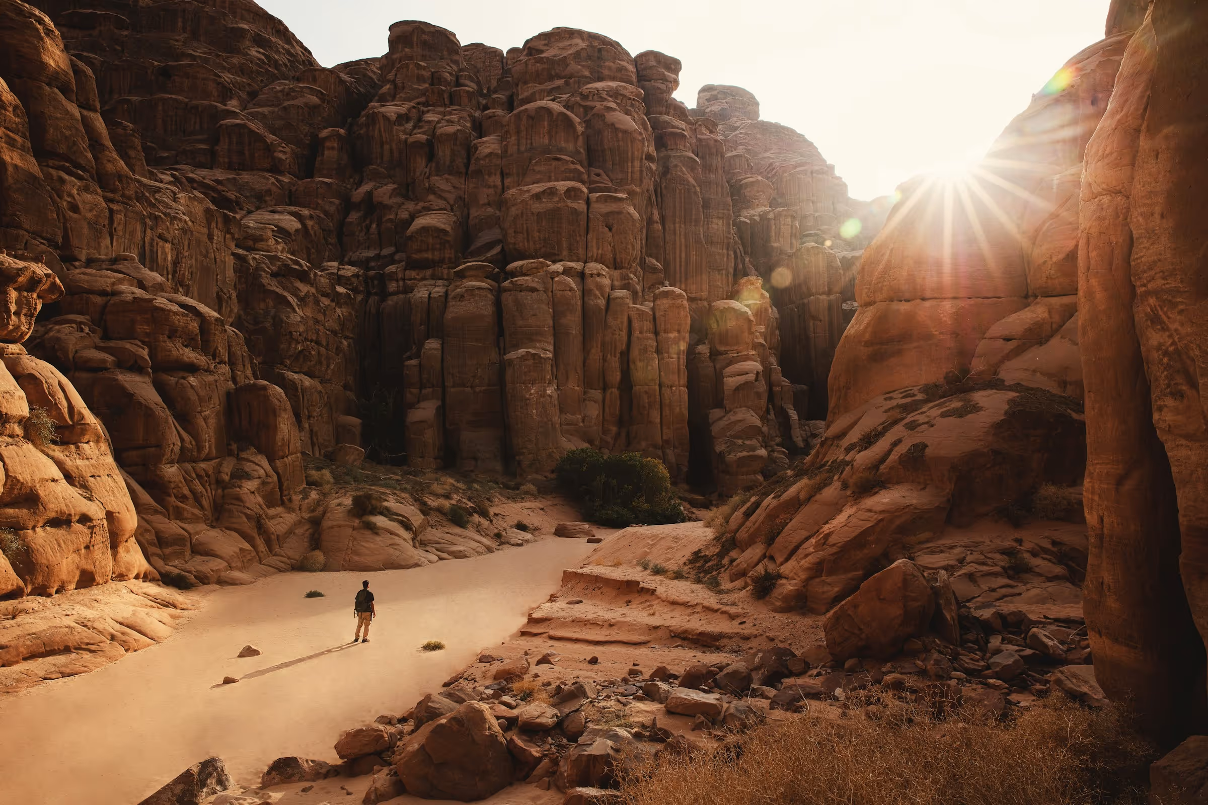 Person standing alone on a sandy path surrounded by towering red rock formations with the sun shining above the rocks.