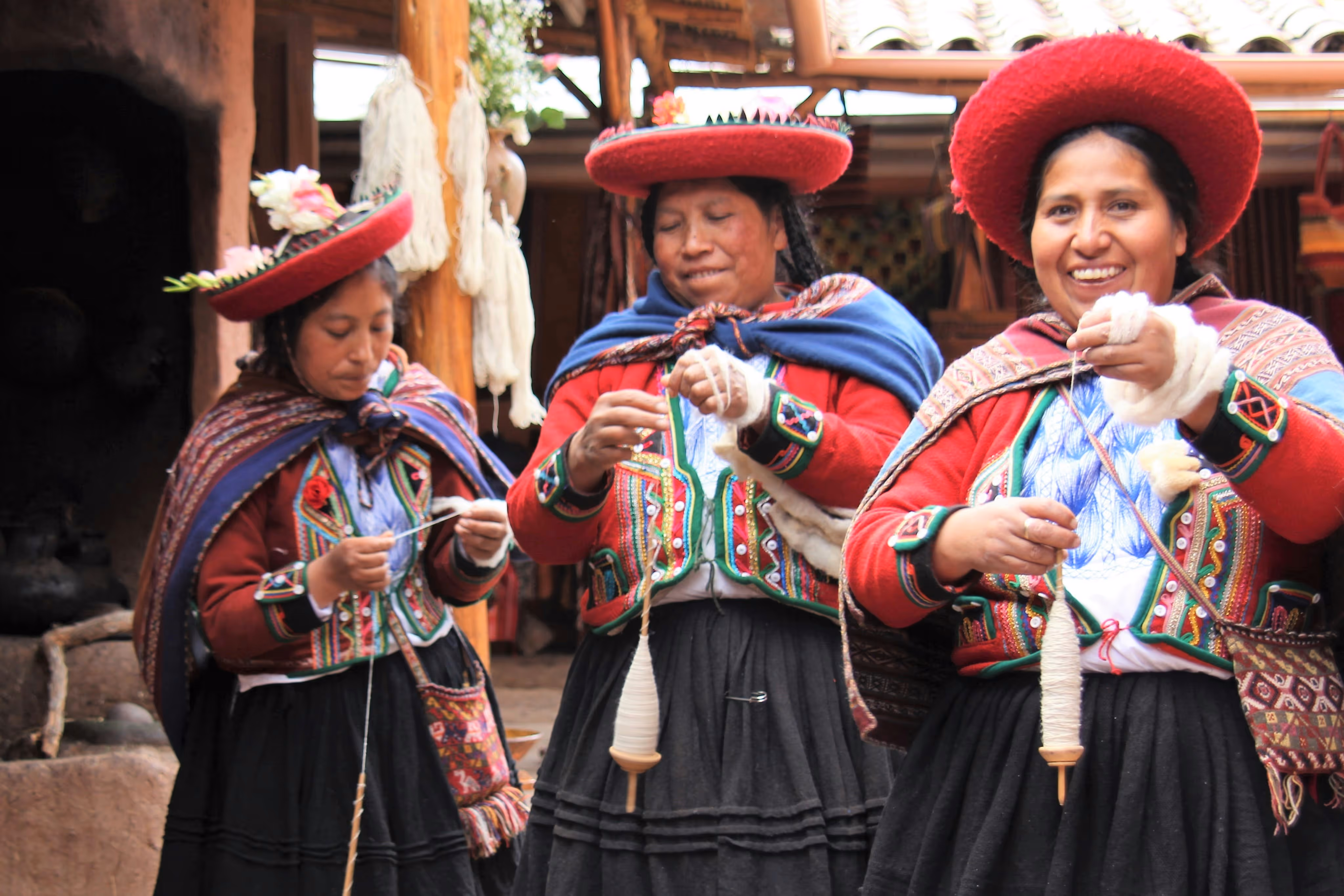 Three indigenous women in traditional colorful clothing spinning wool with drop spindles, two smiling and one focused on her work.