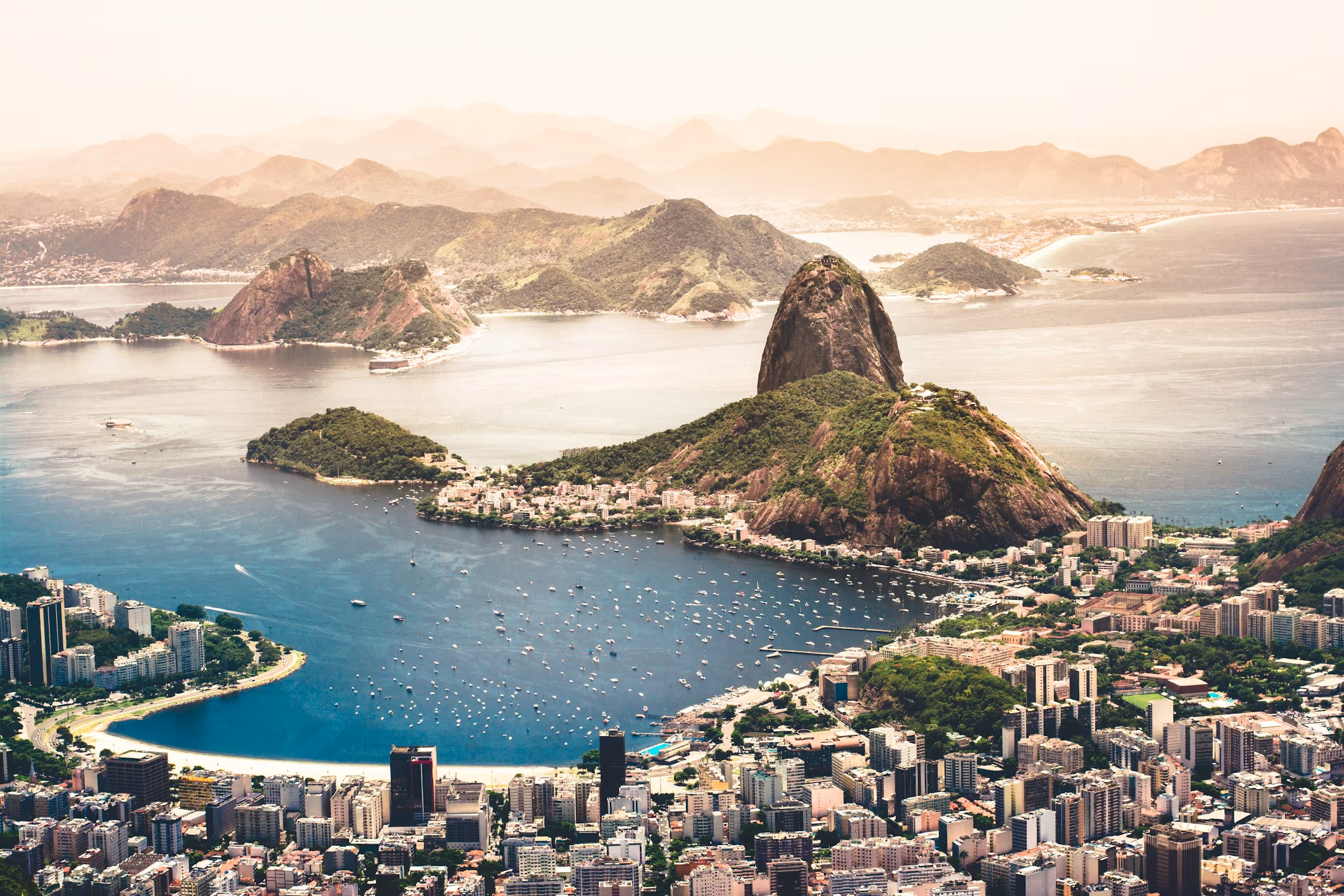 Aerial view of Rio de Janeiro featuring Sugarloaf Mountain, surrounding hills, city buildings, and calm bay with numerous boats.