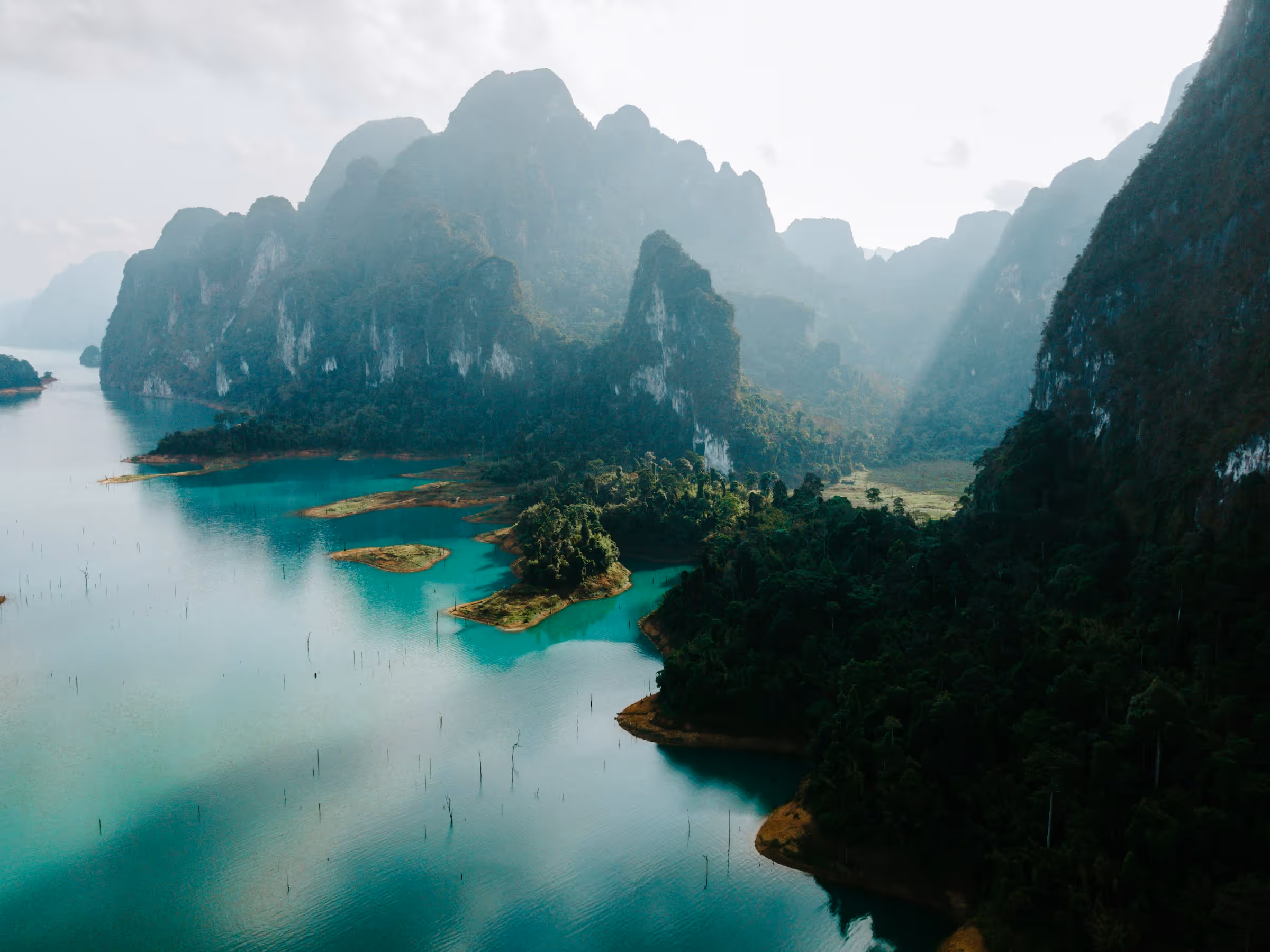 Aerial view of turquoise water surrounded by lush green forest-covered limestone mountains under a misty sky.