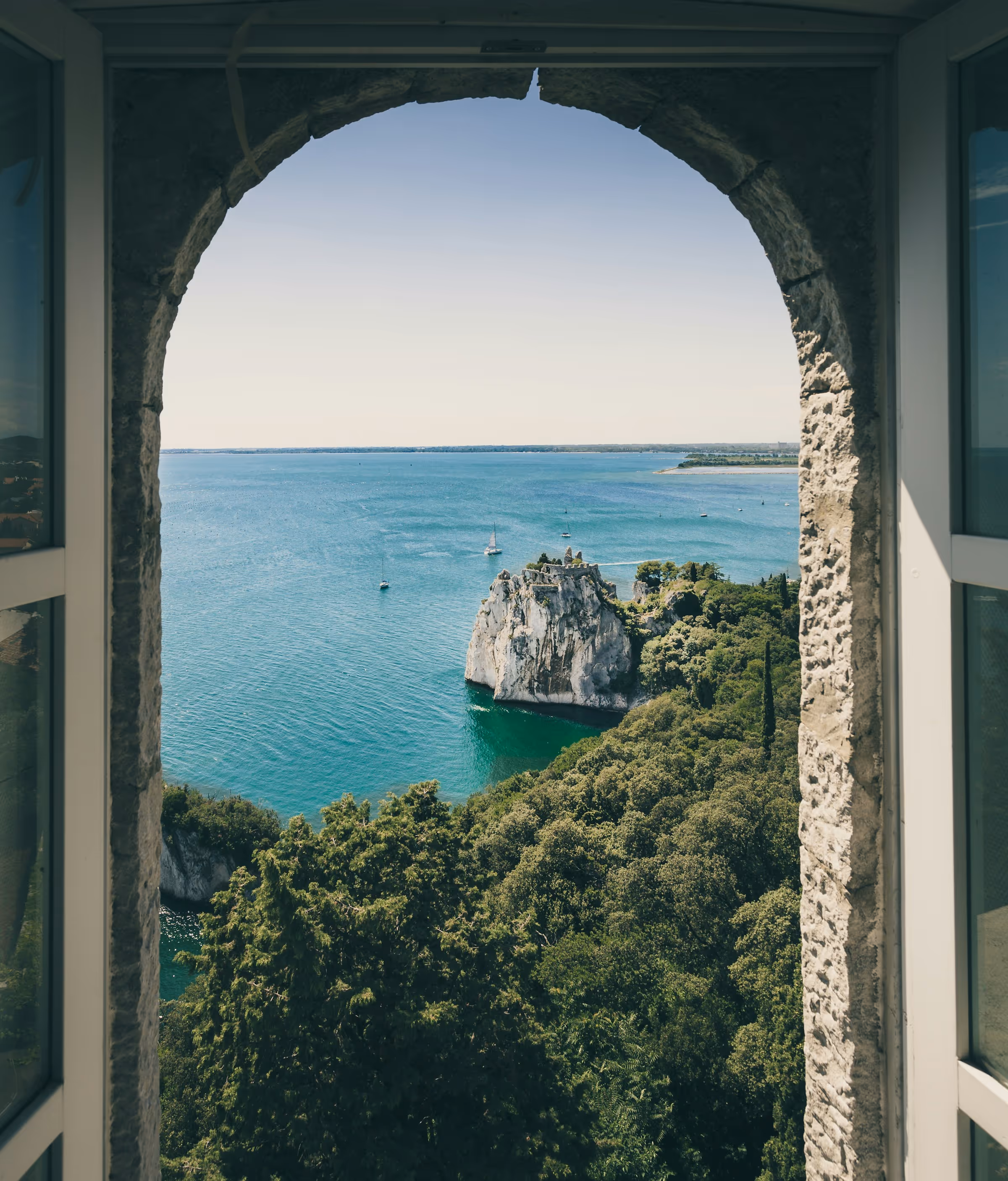 View through a stone archway overlooking blue sea, rocky cliff with ruins, sailboats, and dense green trees.
