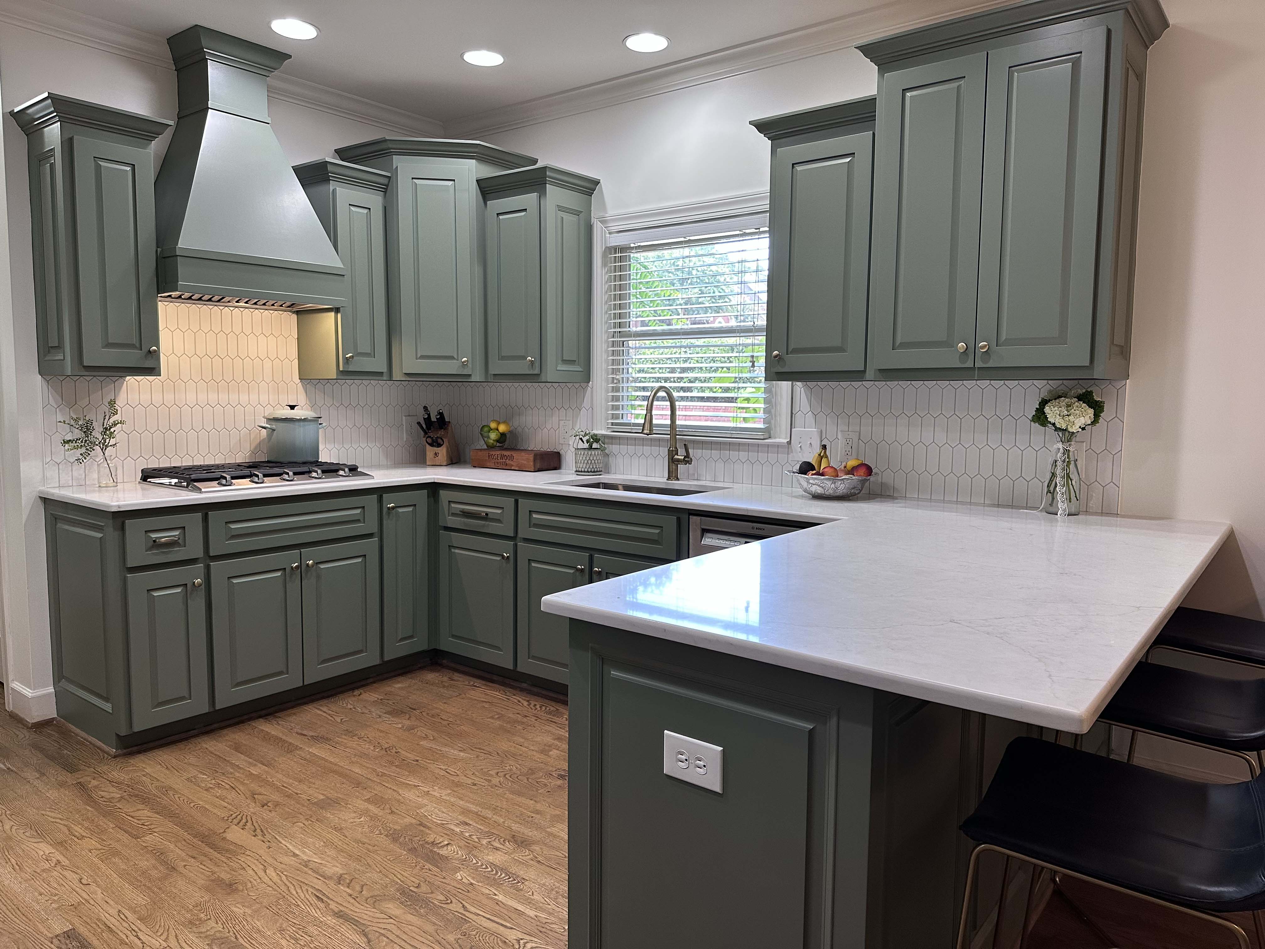 Modern kitchen with green cabinetry, white hexagonal tile backsplash, white marble countertops, stainless steel sink under window, gas stove, and wooden floor.